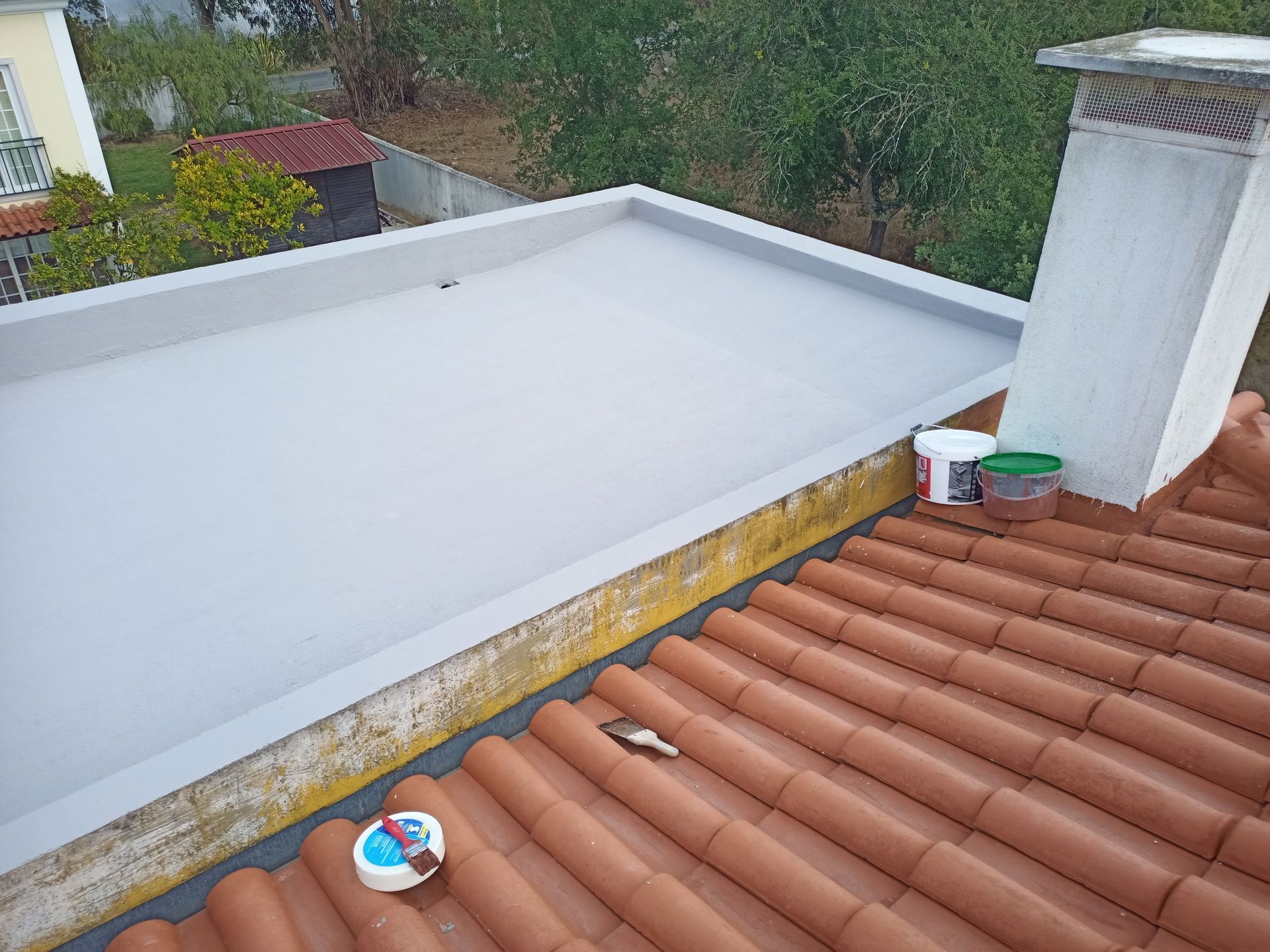 Telhado plano cinza claro adjacente a um telhado de telha vermelha com chaminé e latas de tinta.