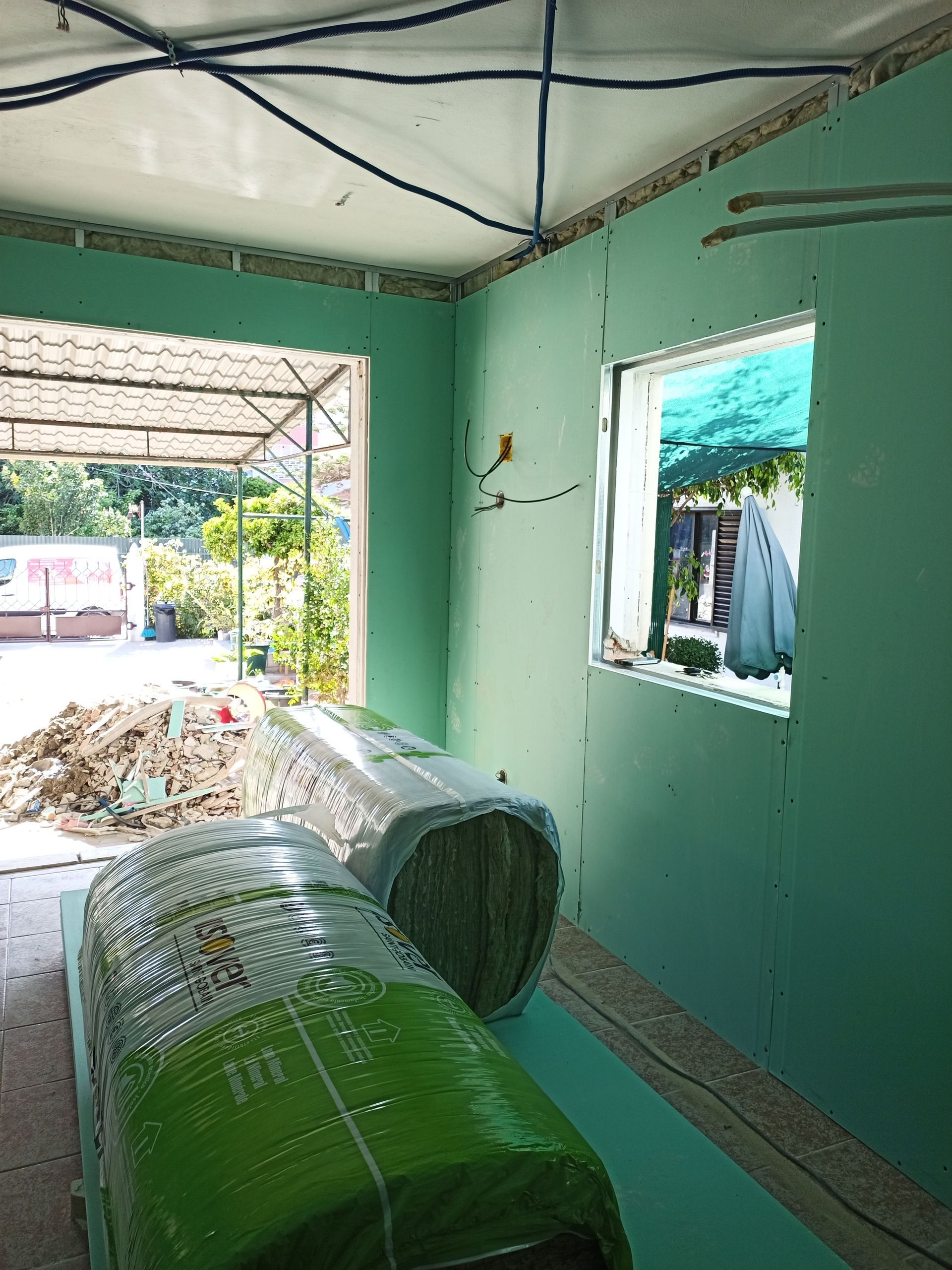 Construção interna com drywall verde, fiação aparente e isolamento em rolo. Vista externa através das janelas.