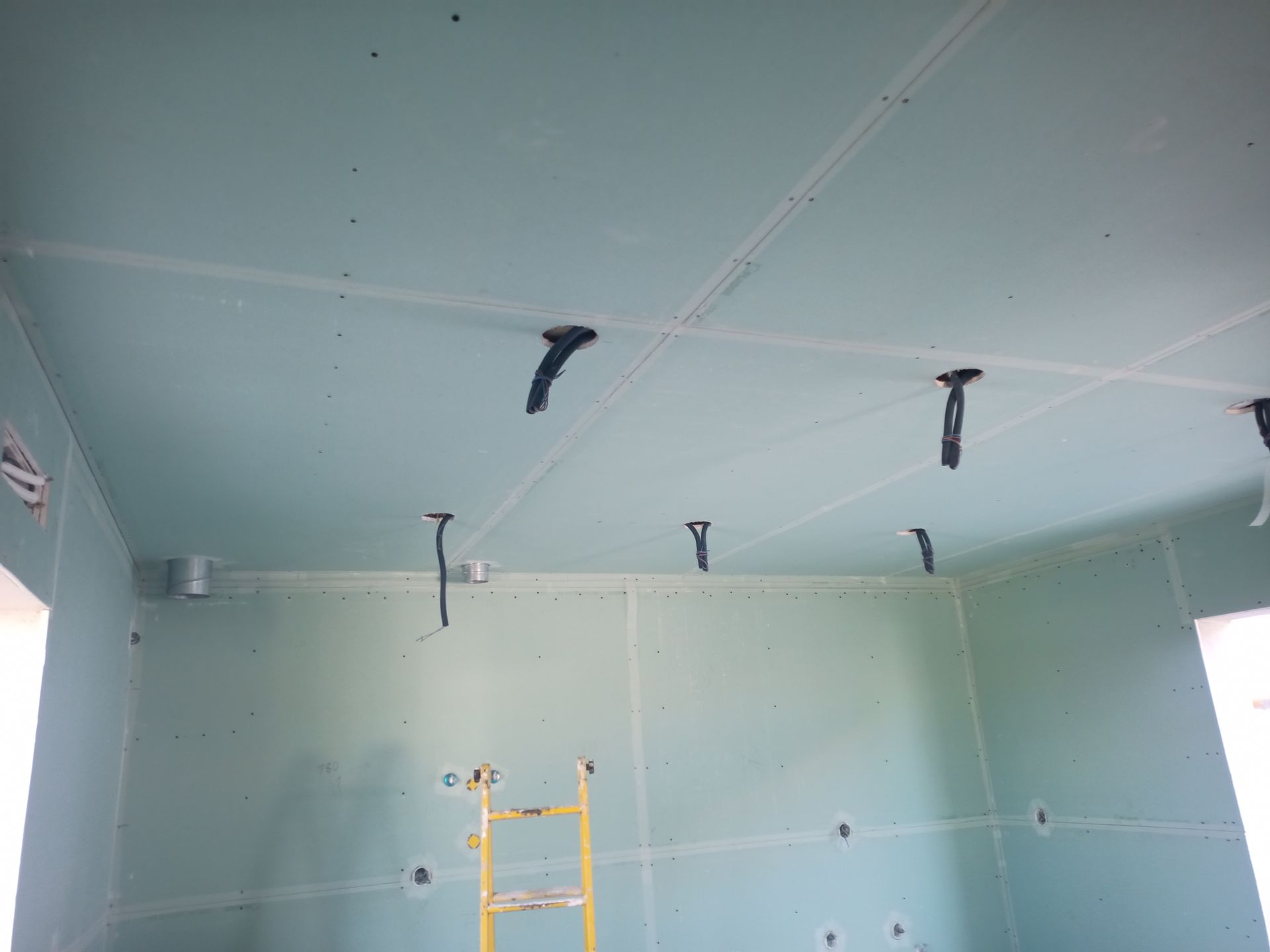 Sala interna em construção, com drywall verde nas paredes e no teto; fios elétricos aparentes.