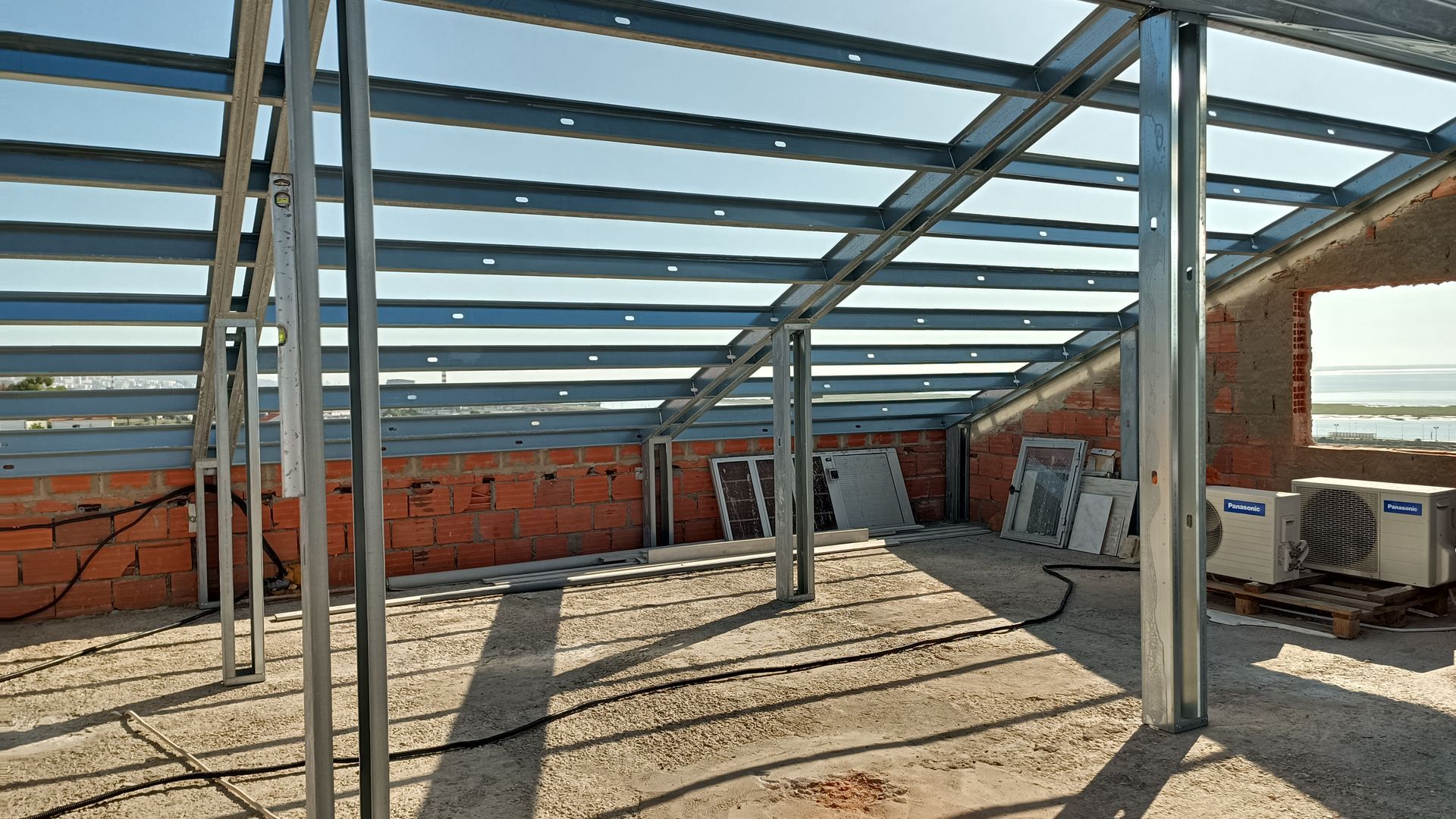 Canteiro de obras em ambiente interno com estrutura metálica para telhado e paredes; tijolos vermelhos visíveis.