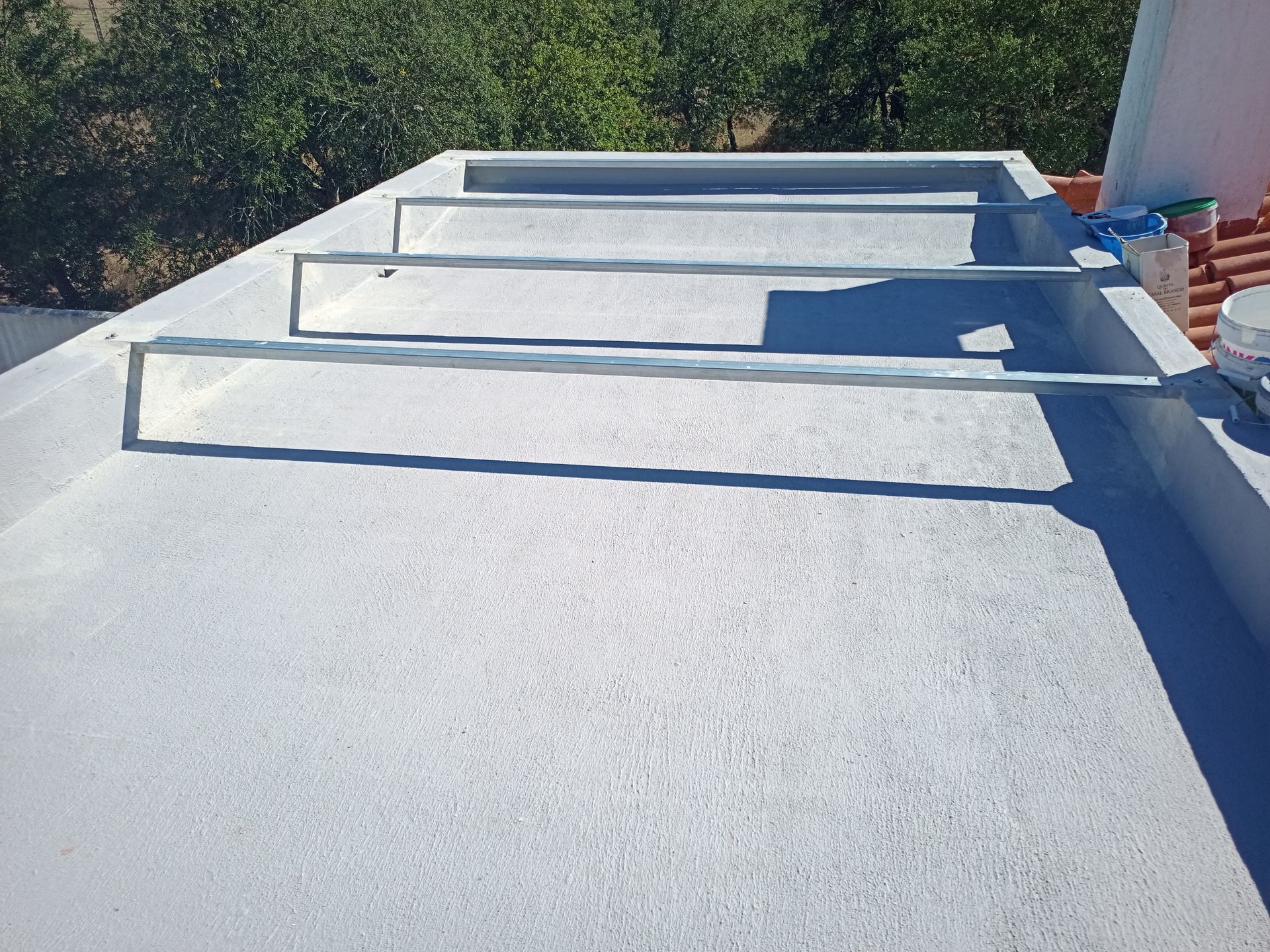 Telhado cinza com barras metálicas horizontais, possivelmente de uma pérgola, com uma fileira de árvores ao fundo.