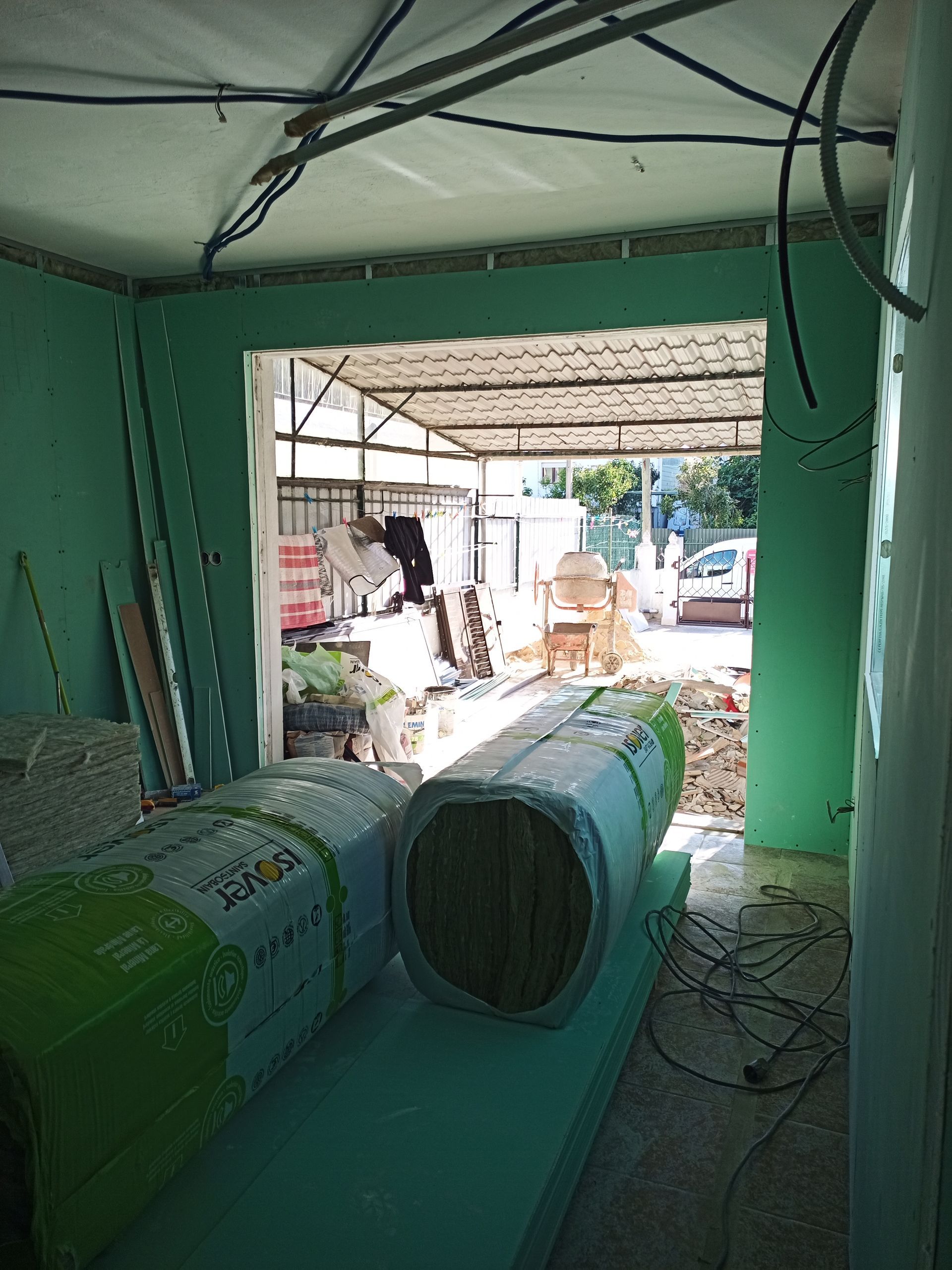 Vista interna de um canteiro de obras com paredes verdes, porta aberta e rolos de isolamento.