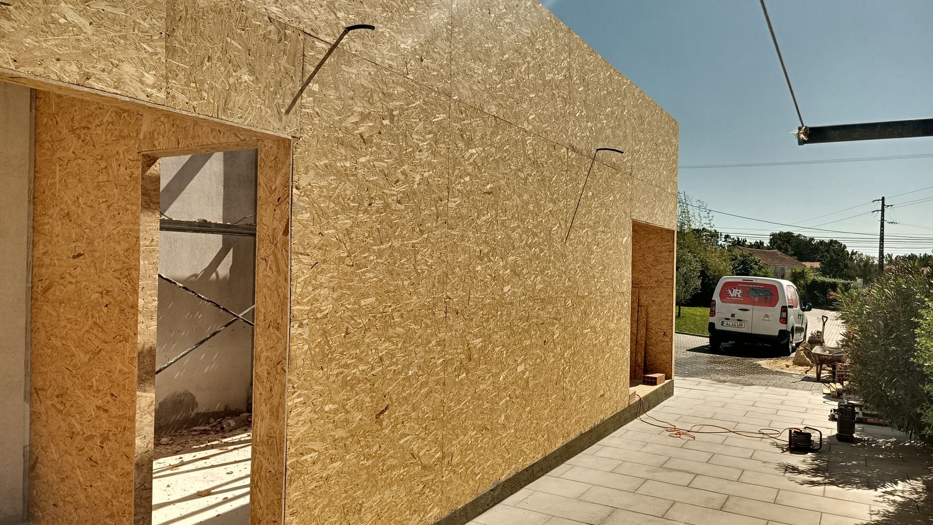 Exterior de um edifício em construção, paredes de painéis OSB (Oriented Strand Board), aberturas de portas e janelas, dia ensolarado.