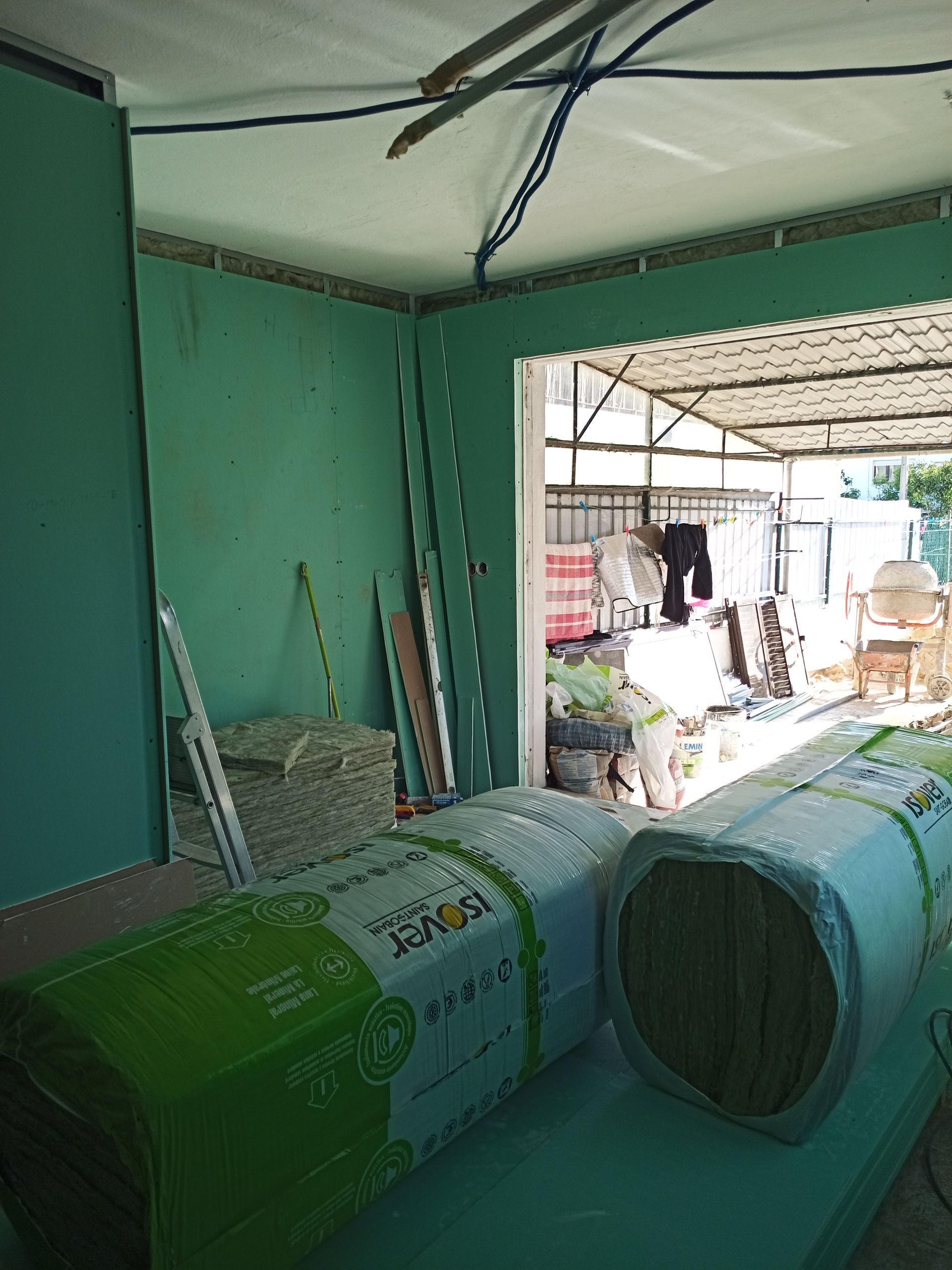 Cena de construção interna com drywall verde, rolos de isolamento e uma porta que dá para o exterior.