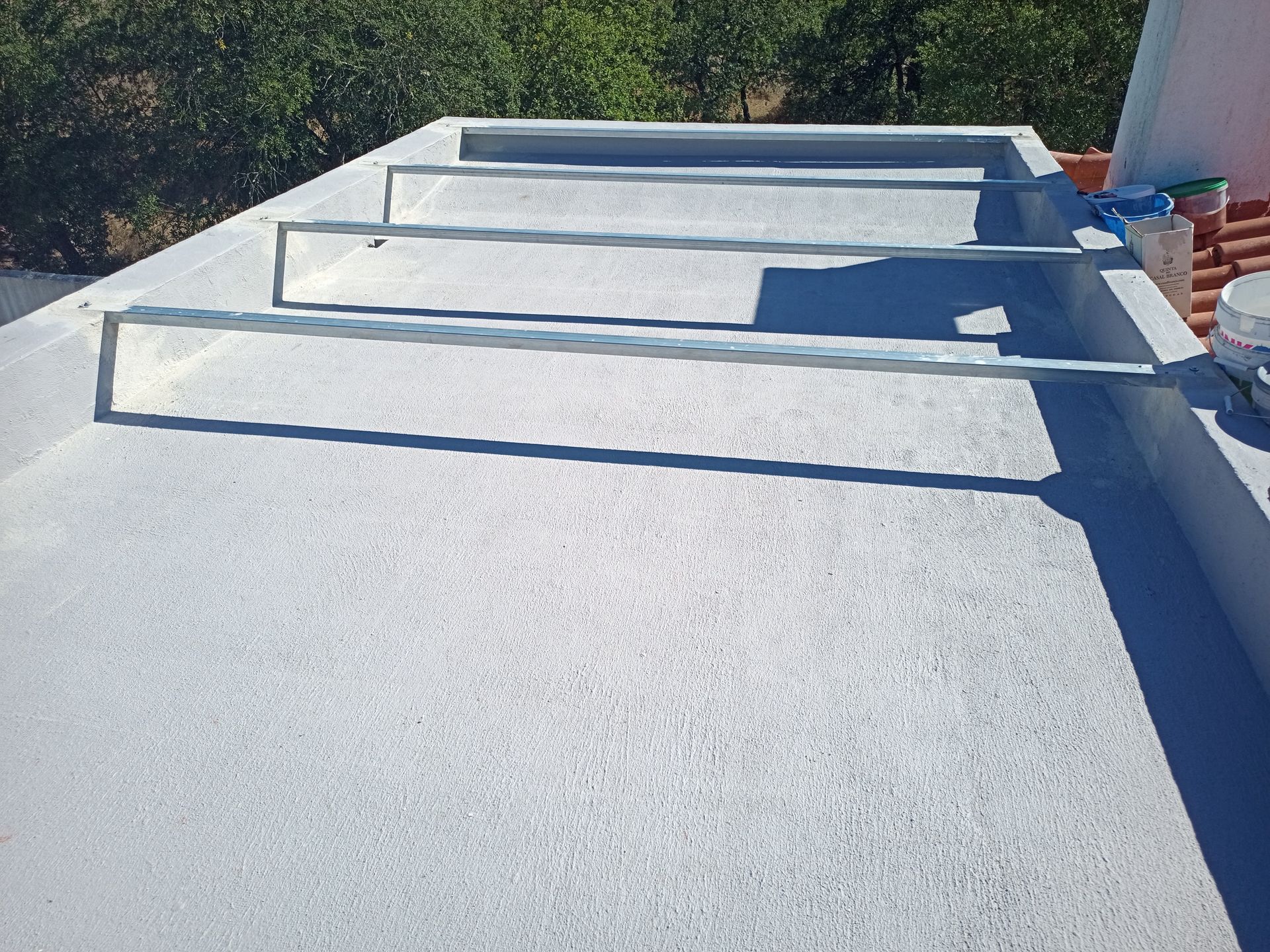 Telhado plano cinza com suportes metálicos; a luz do sol projeta sombras.