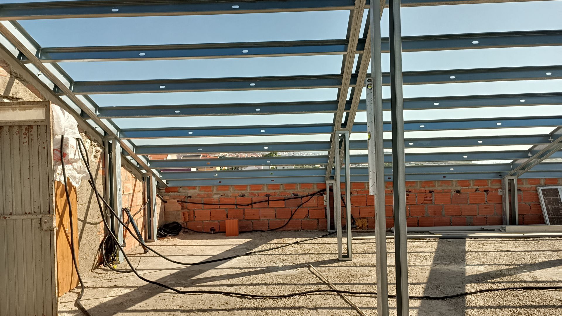 Canteiro de obras: estrutura metálica de um telhado sobre paredes de tijolo; fios e entulho no chão.