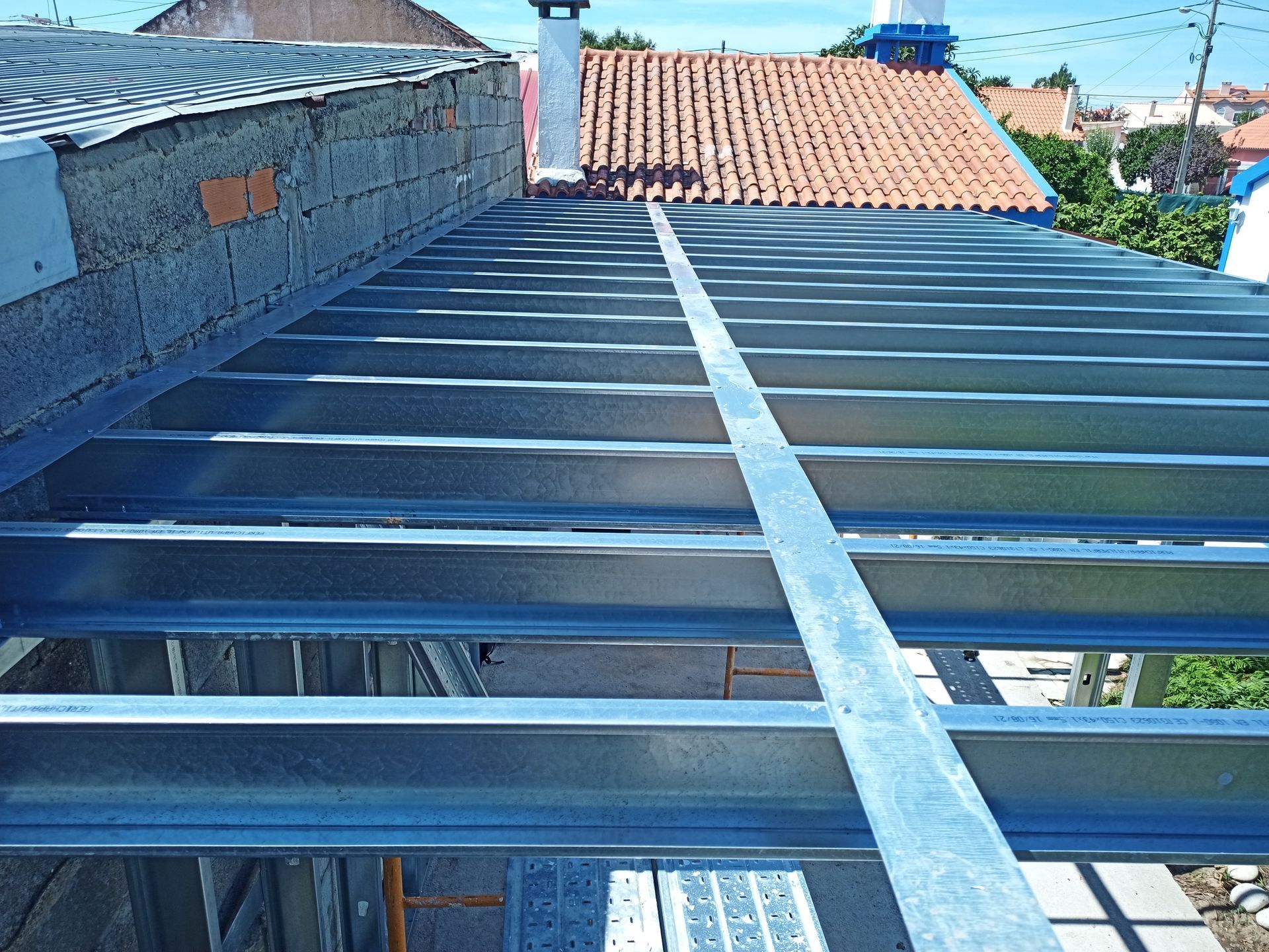 Estrutura metálica de um telhado em construção, com um telhado de telhas vermelhas ao fundo.