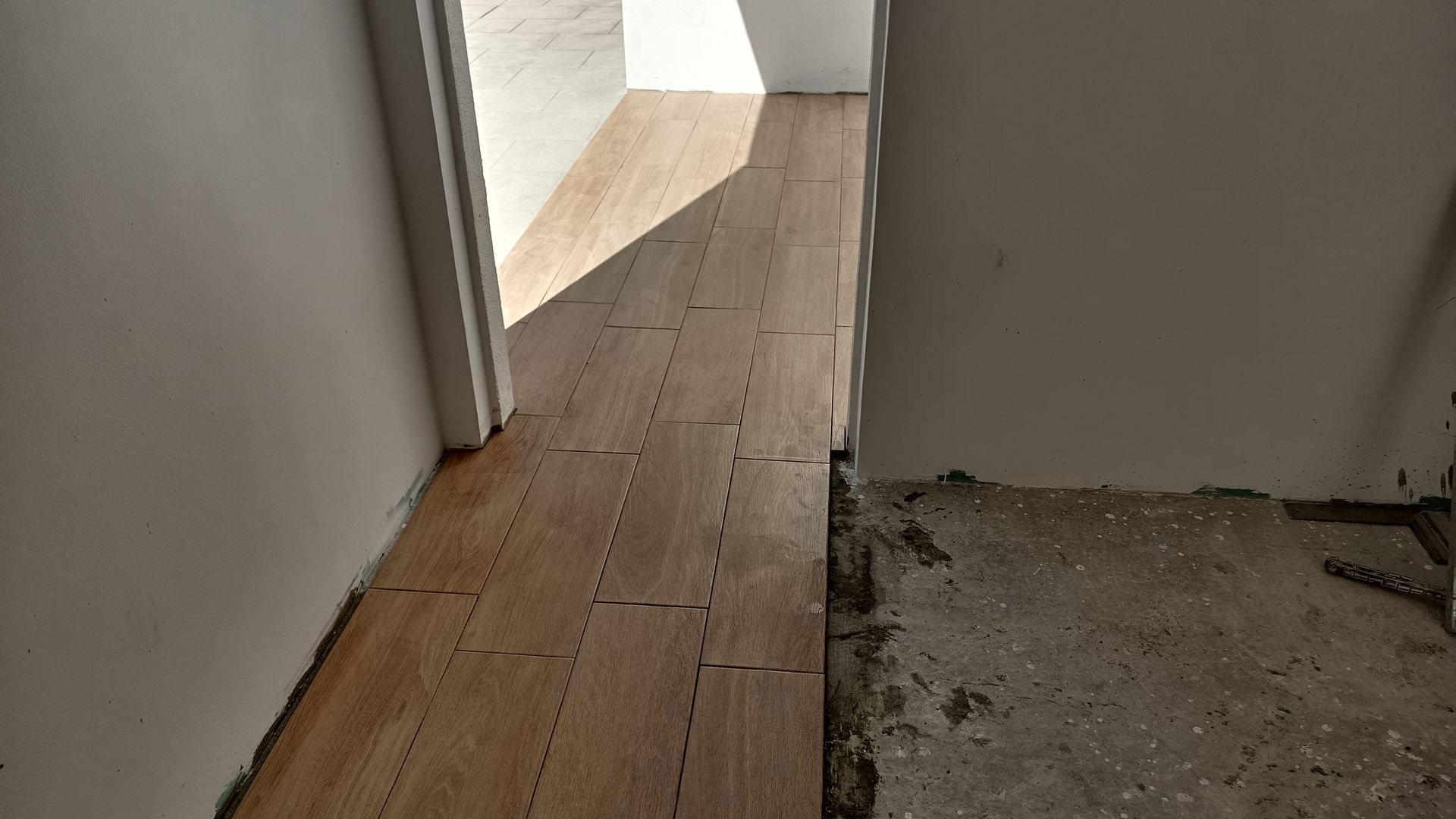 Corredor com piso de madeira novo à esquerda e concreto inacabado à direita. Porta no final.