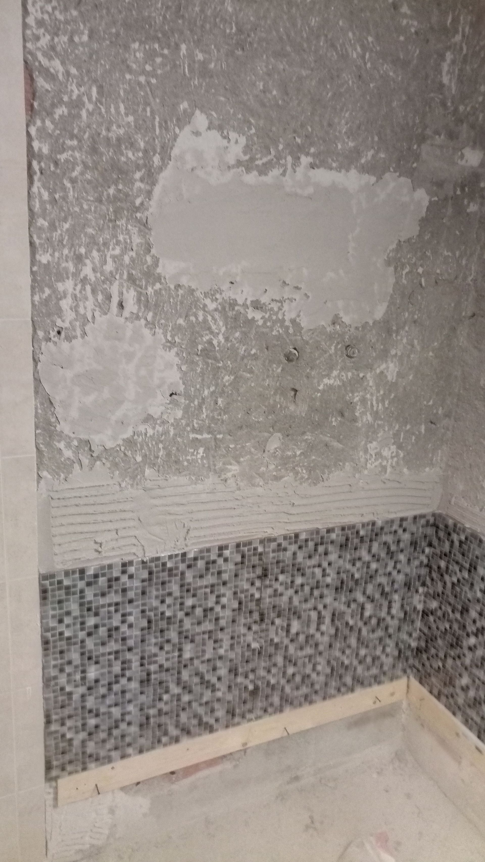 Parede do banheiro em construção, base de azulejo mosaico, concreto aparente acima.