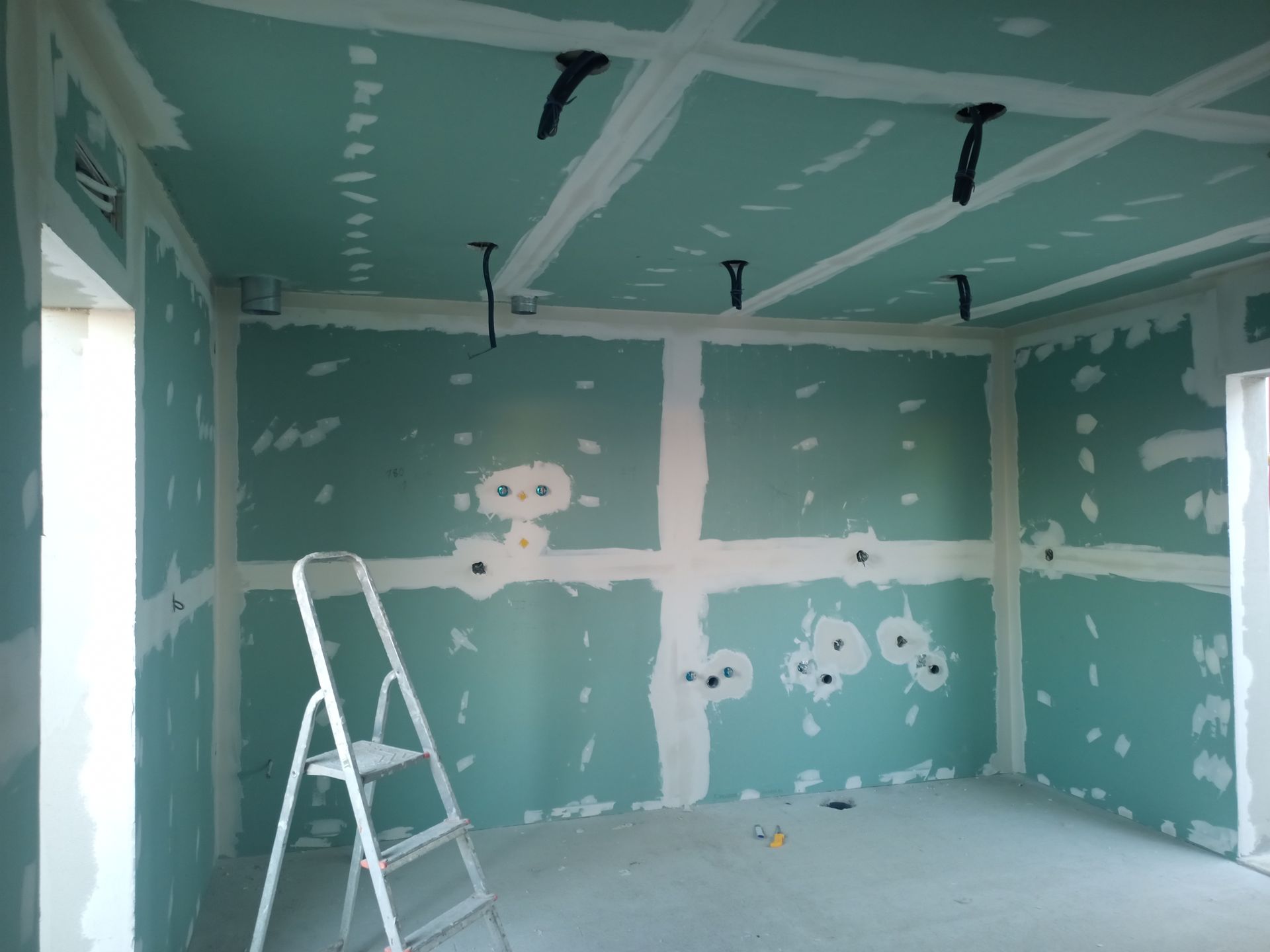 Construção interna: Sala com drywall verde e massa corrida branca, uma escada e conduíte elétrico.