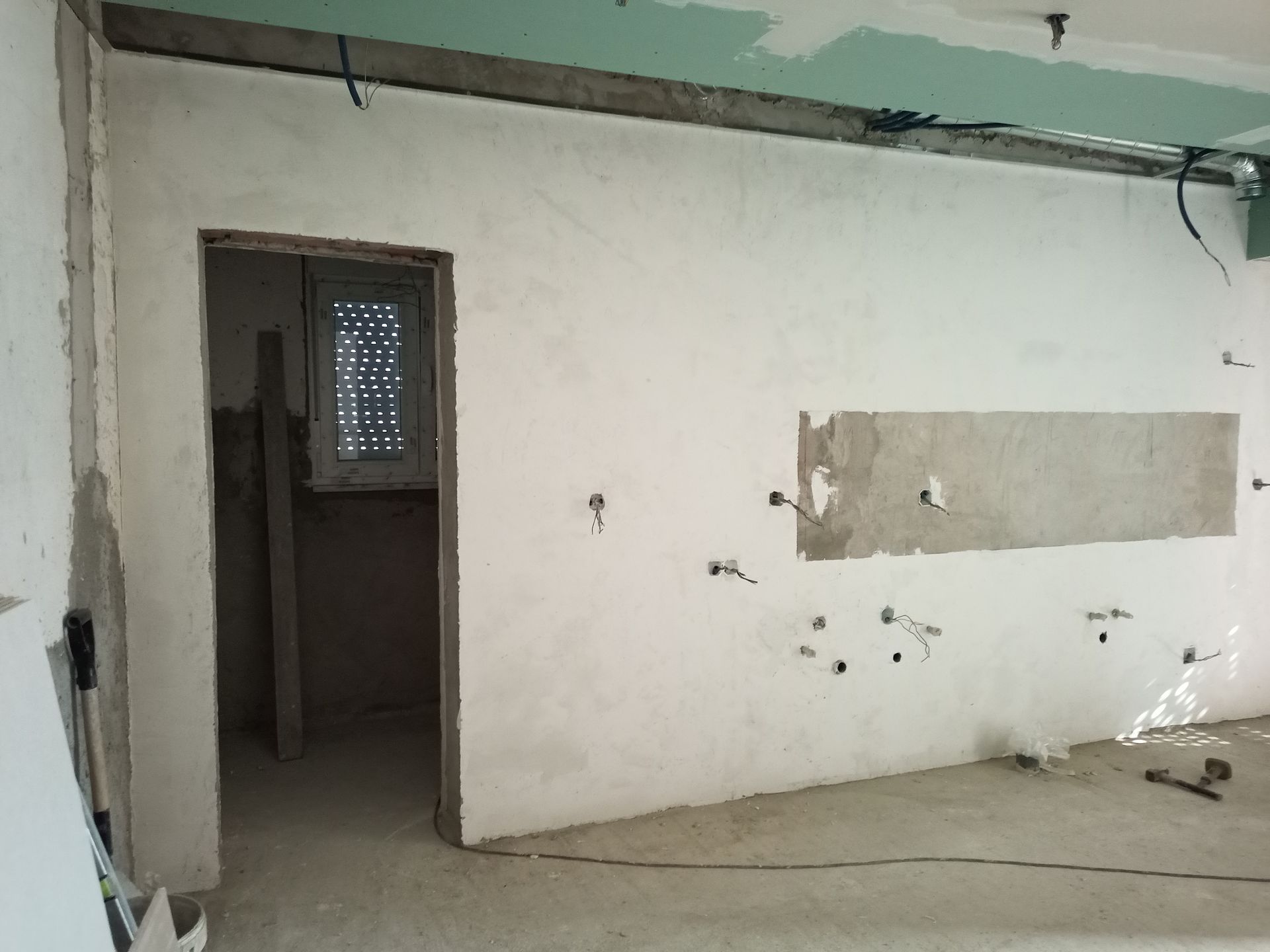 Interior em construção: paredes brancas com tomadas elétricas, porta, vigas expostas no teto e uma pequena janela.