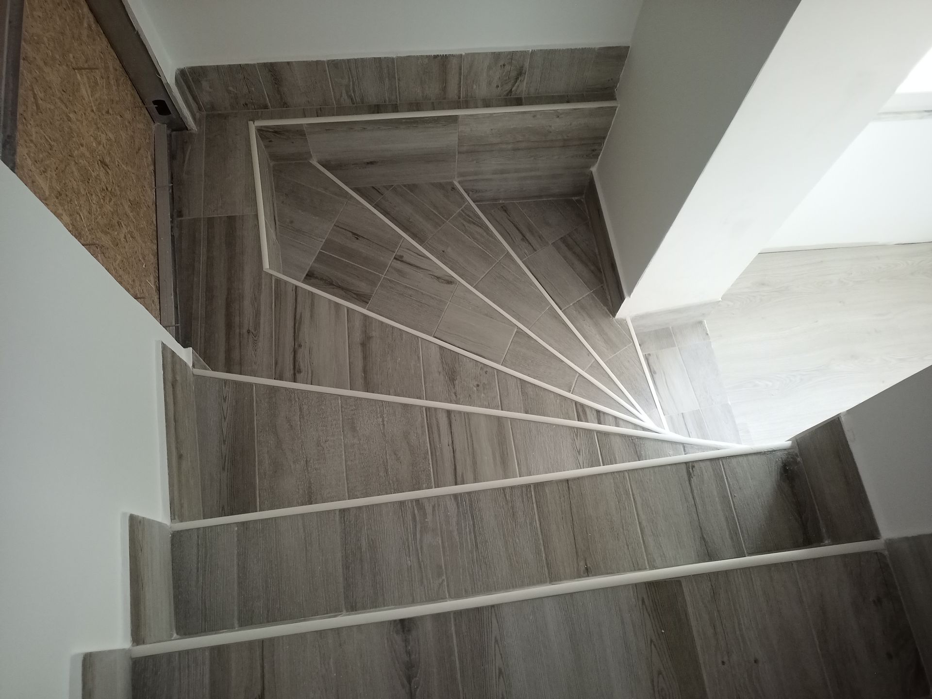 Vista de cima para baixo de uma escada moderna revestida com azulejos que imitam madeira e com detalhes em branco, que leva ao andar inferior.