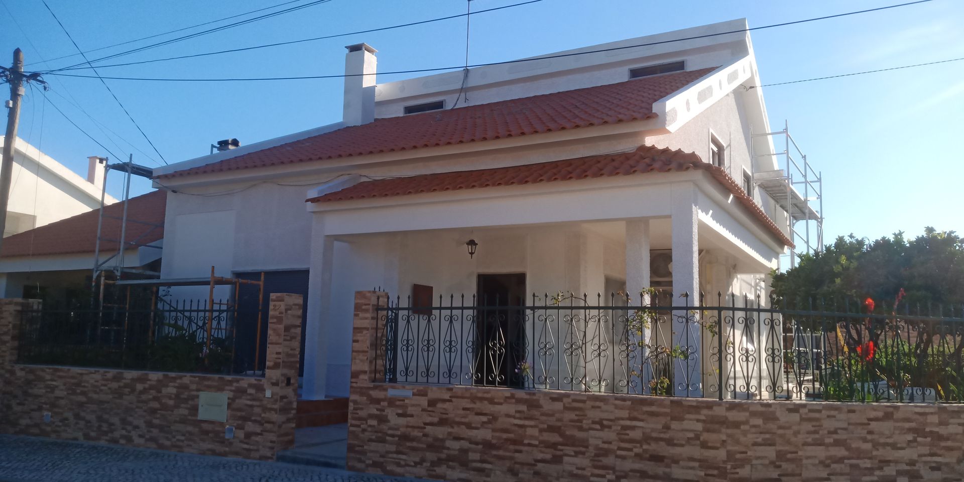 Uma casa de dois andares com telhado de telhas vermelhas, paredes brancas e varanda, atrás de um muro de tijolos e uma cerca de metal preta.