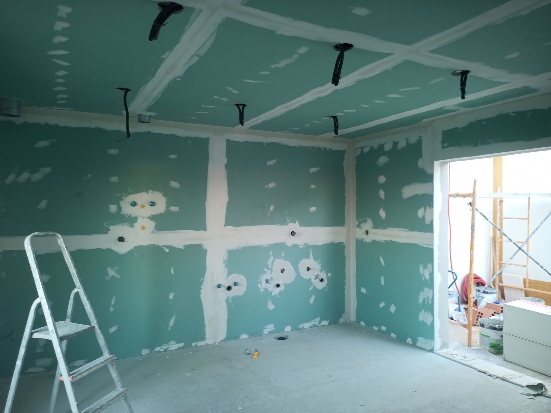 Sala em construção com drywall verde e massa corrida branca. Uma escada está encostada na parede.