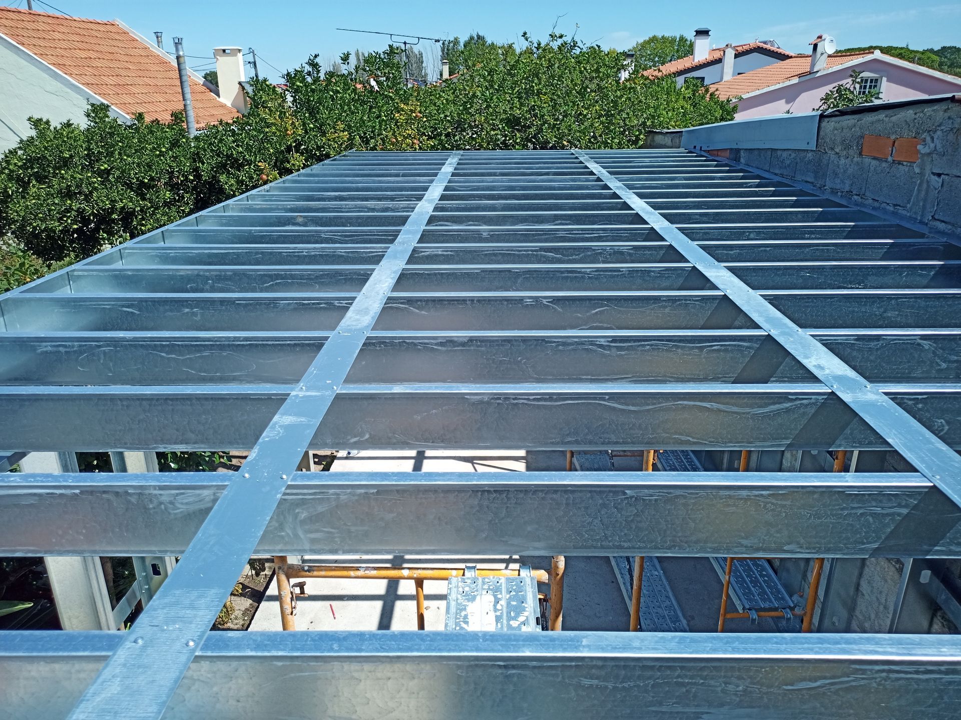 Estrutura metálica de um telhado em construção, tendo como pano de fundo árvores e casas.