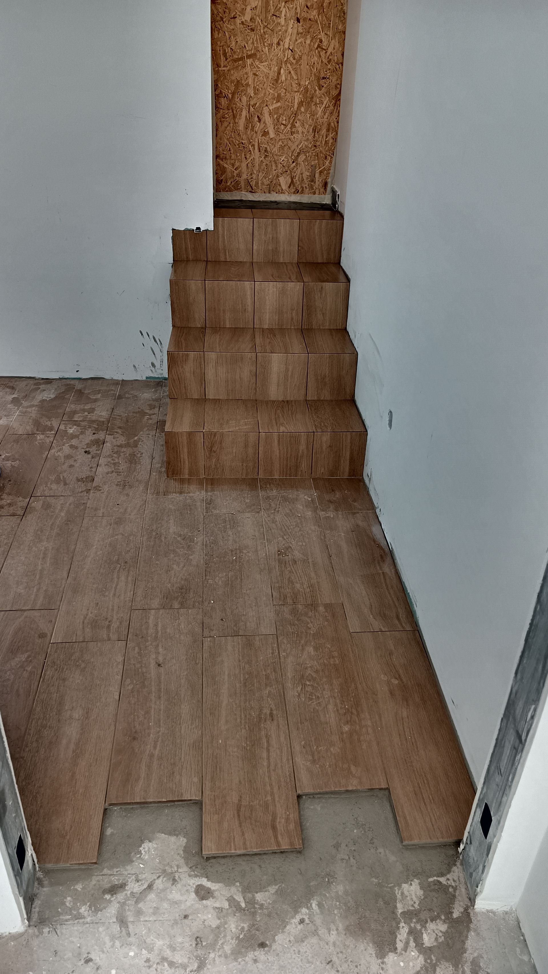 Escada de madeira que leva a um andar superior a partir de um cômodo com piso inacabado e paredes brancas.