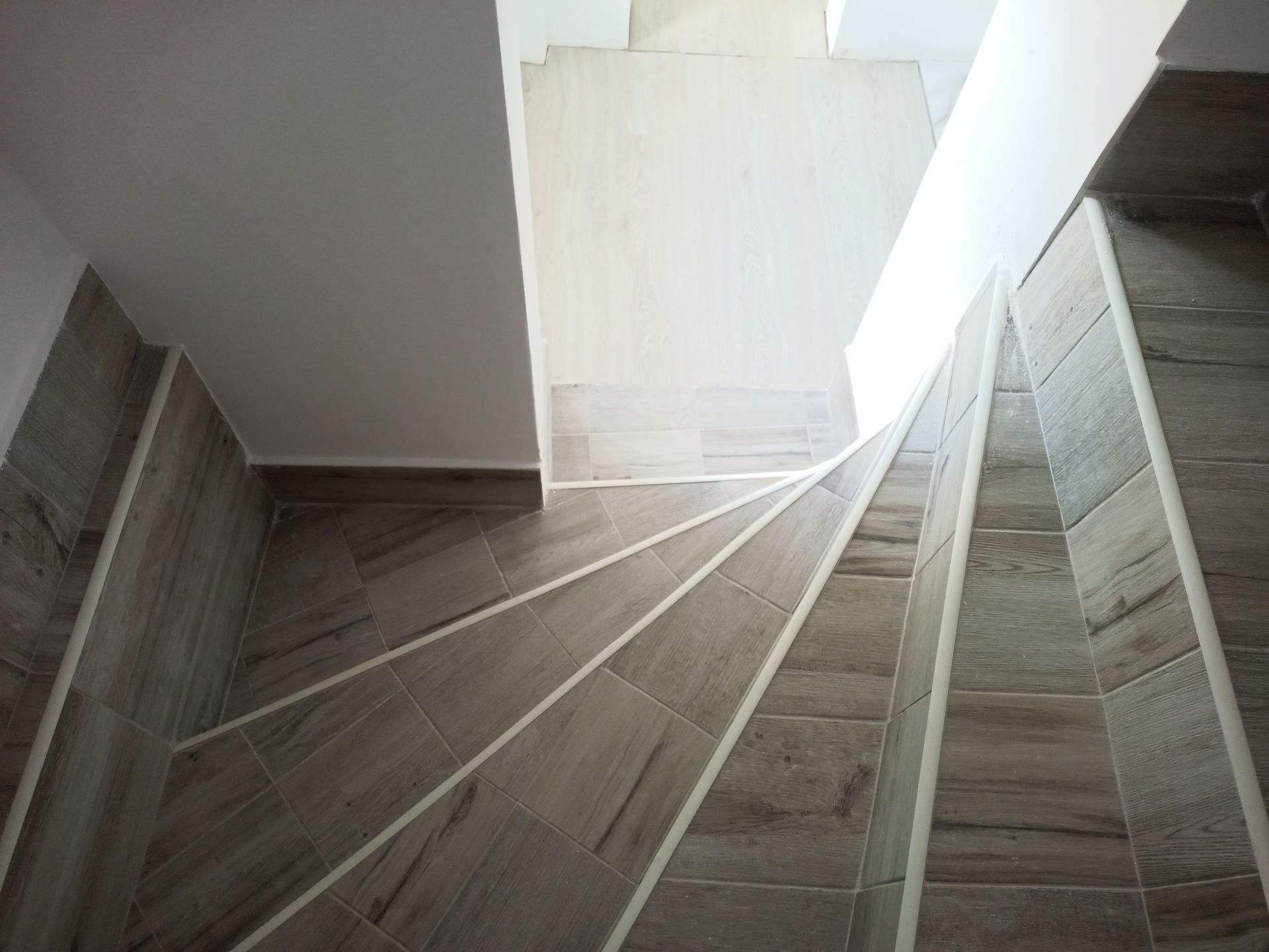 Vista aérea de uma escada em espiral com degraus revestidos com azulejos que imitam madeira e paredes brancas.