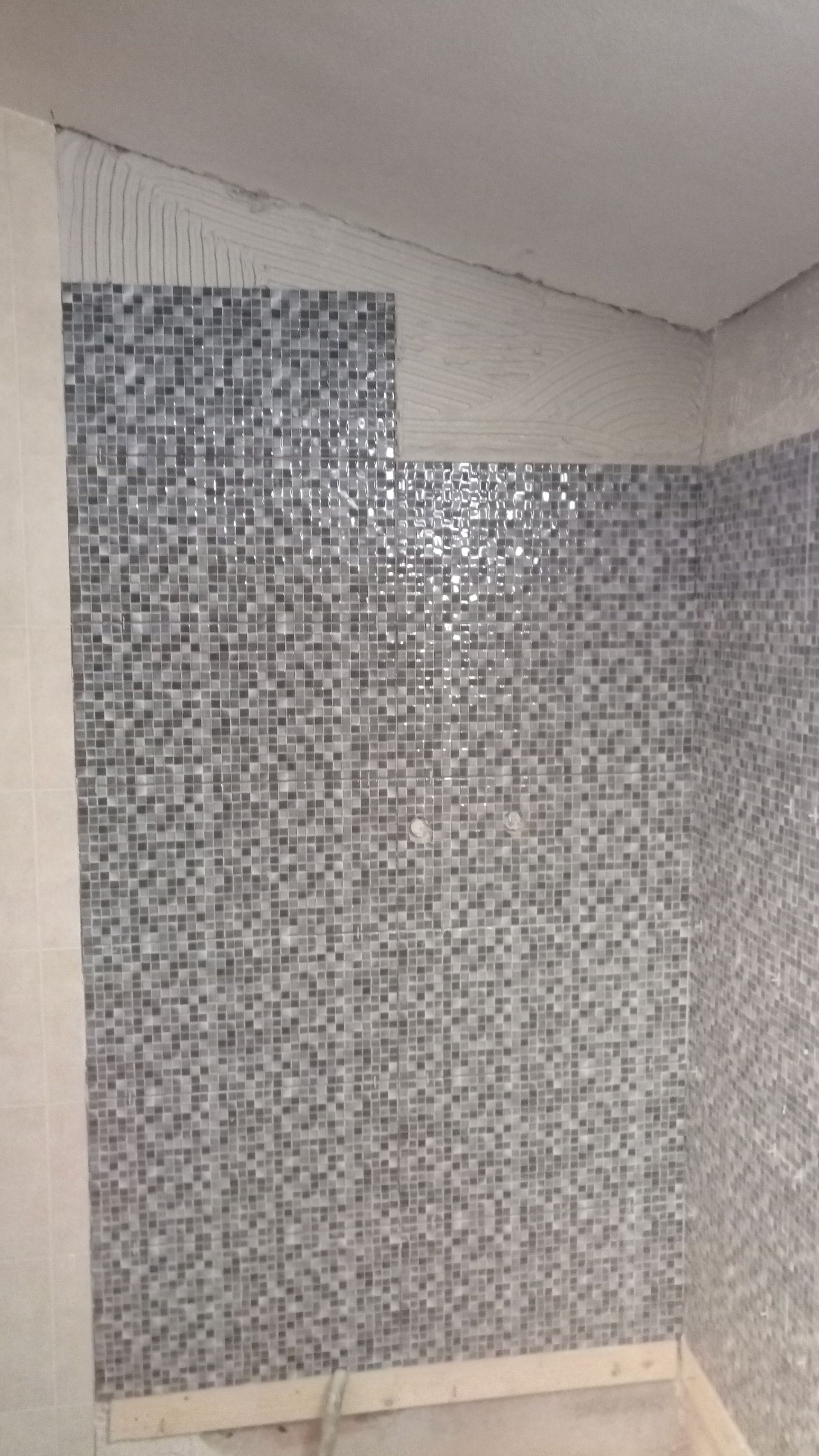 Instalação parcial de mosaico na parede. Predominantemente azulejos prateados, cinza e brancos.