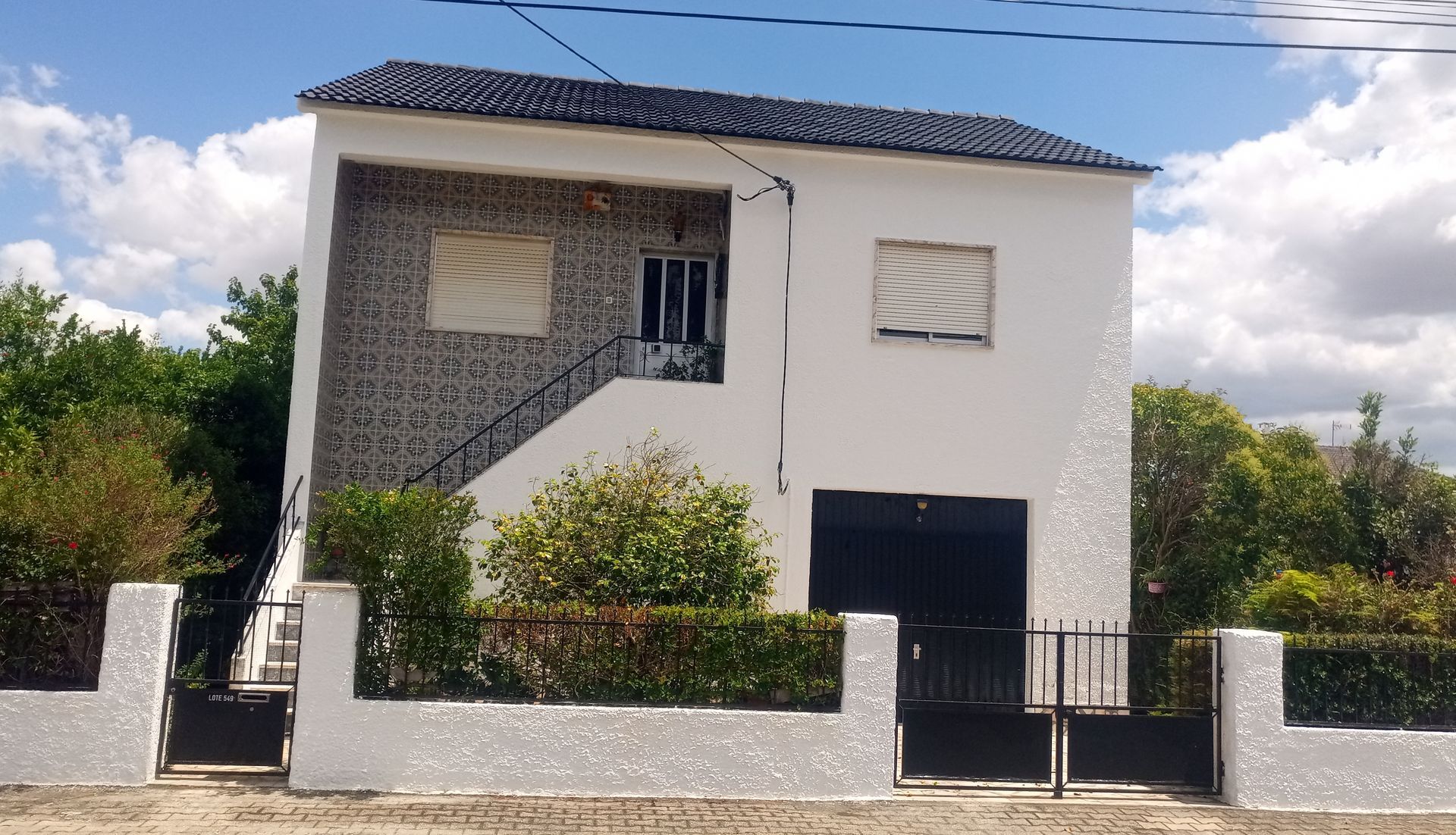 Casa branca de dois andares com telhado e venezianas pretas, cerca preta e arbustos na frente.