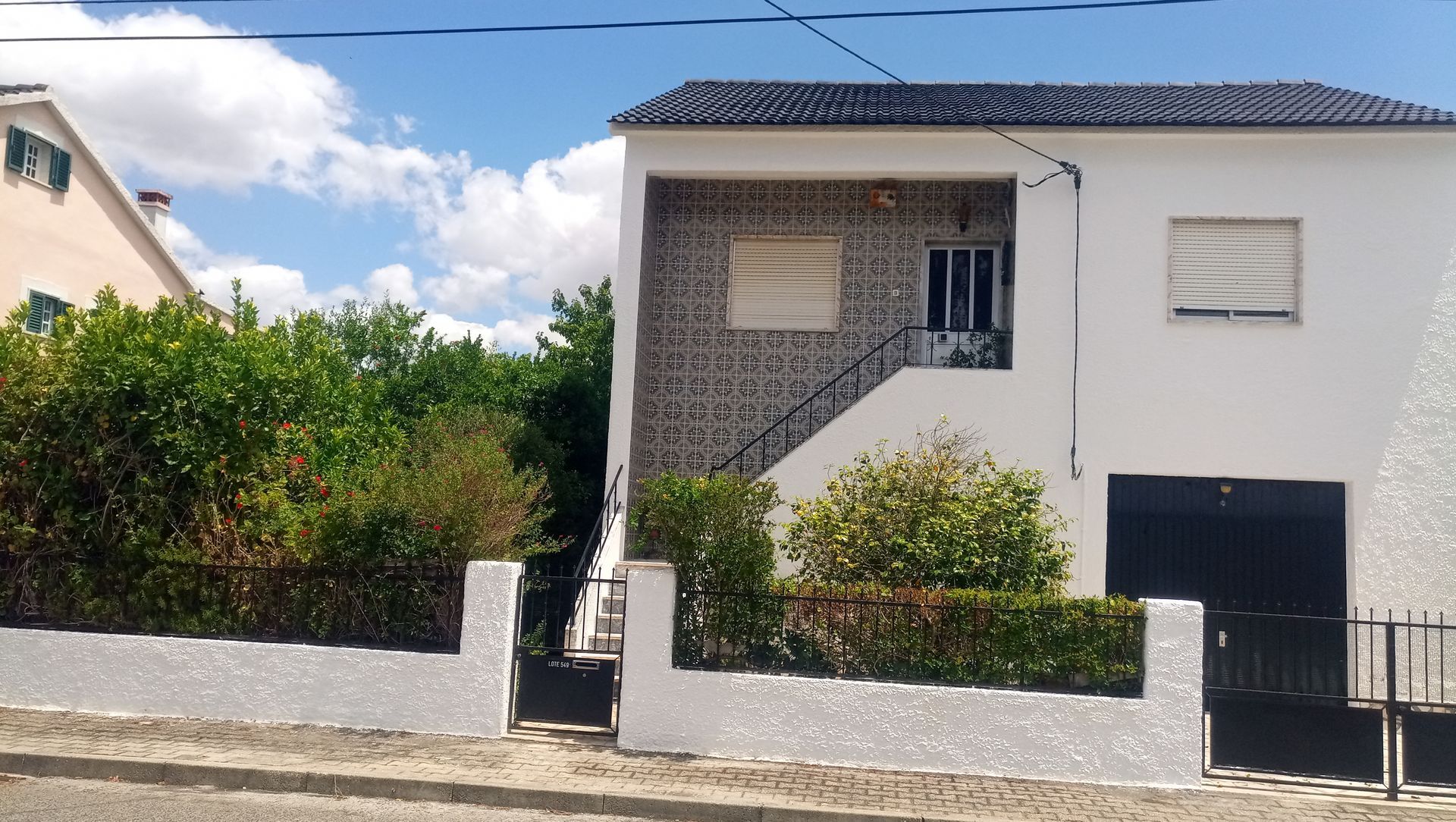 Casa branca de dois andares com porta e garagem pretas, com um muro baixo e vegetação na frente, sob um céu azul.