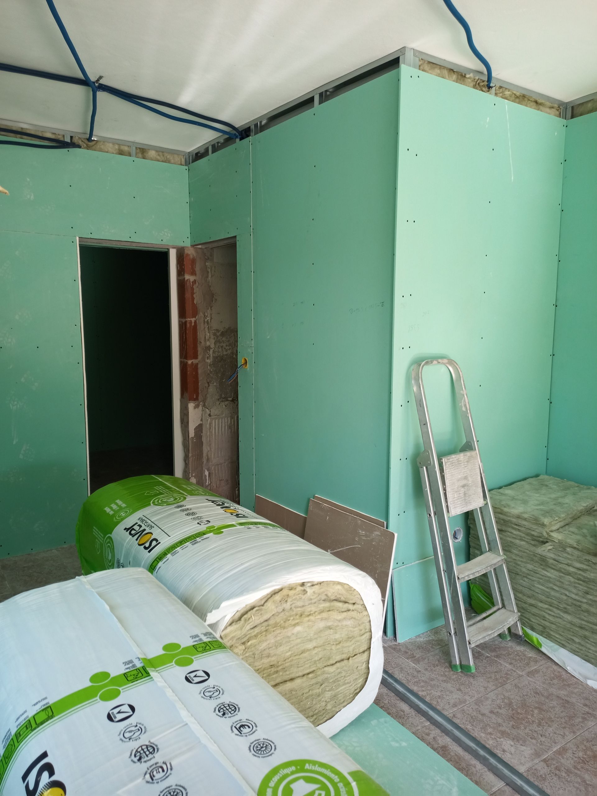 Construção interna com drywall verde, rolos de isolamento, porta e escada.
