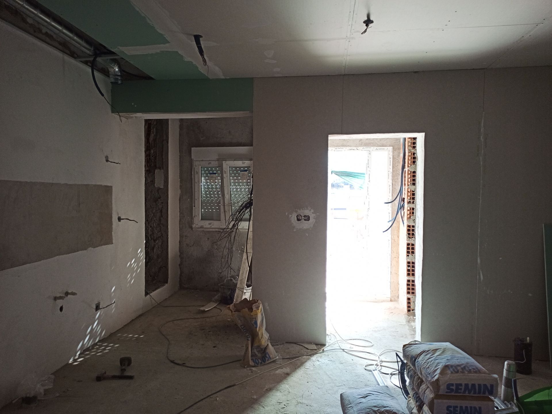 Interior de um cômodo em construção, com paredes de gesso, fiação exposta e luz solar intensa visível através de uma porta.