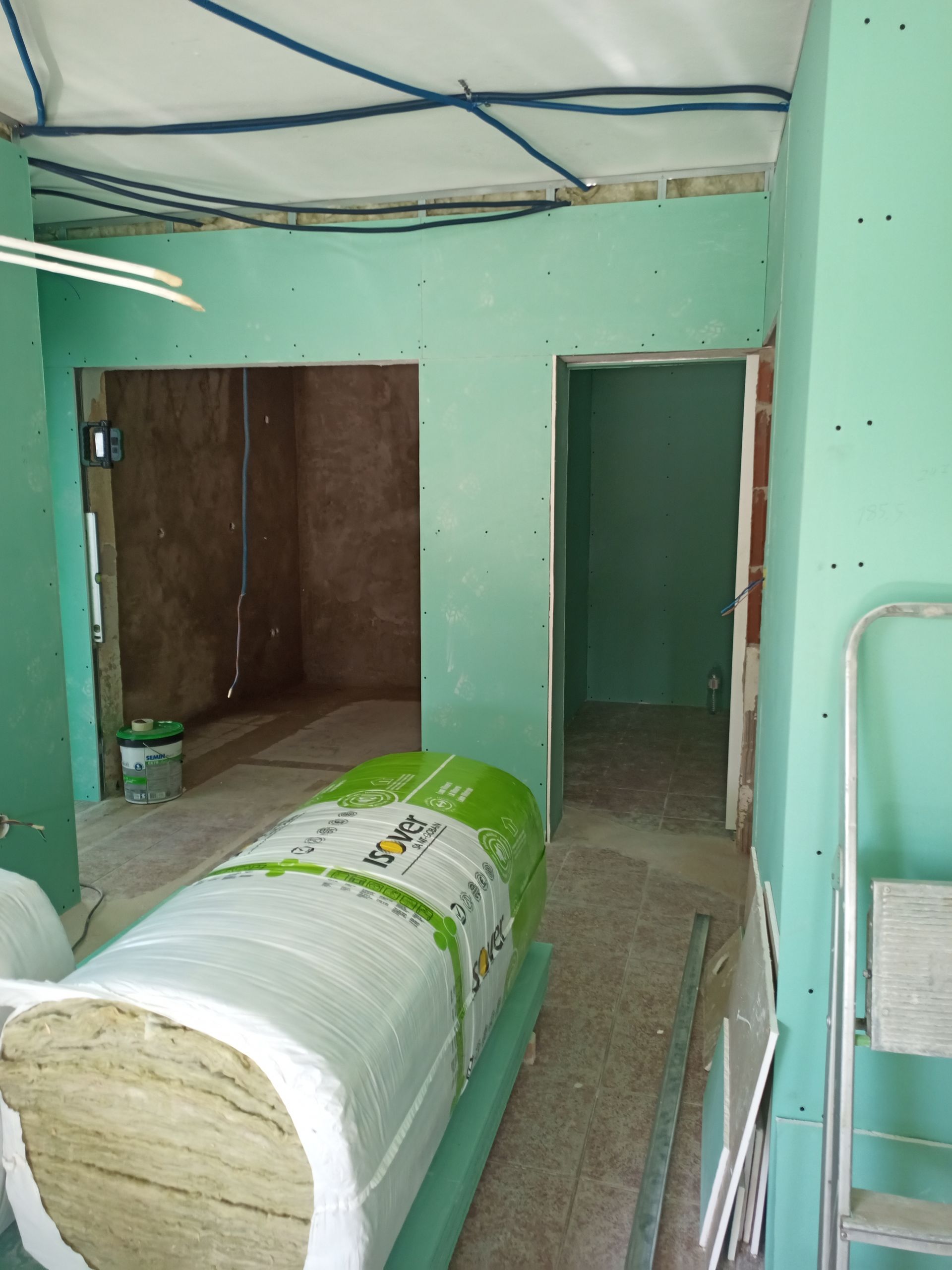 Canteiro de obras interno, drywall verde, rolo de isolamento, portas, fiação azul, escada.