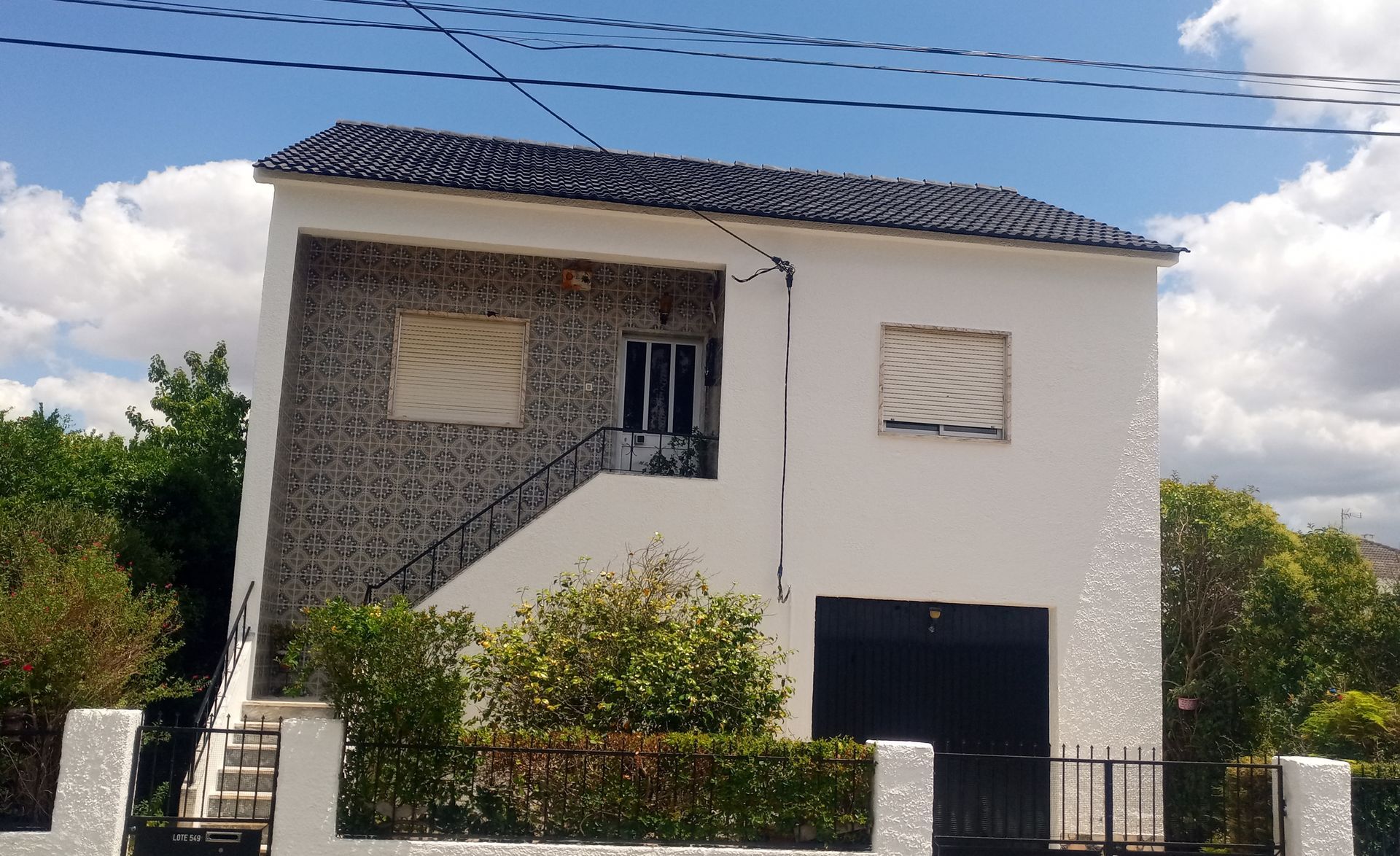 Casa branca de dois andares com telhado escuro e garagem. Azulejos decorativos na varanda. Corrimão e cerca de metal.