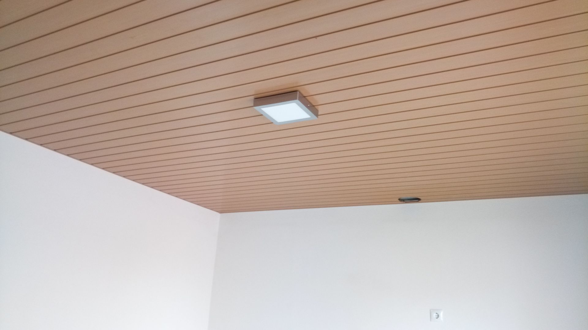 Um quarto com teto ripado de madeira, paredes brancas e uma luminária retangular.