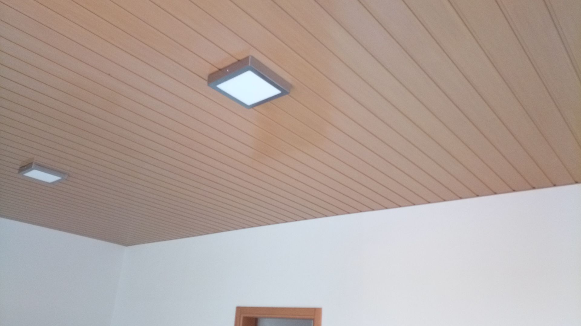 Teto com painéis de madeira e duas luminárias quadradas brancas; paredes brancas.