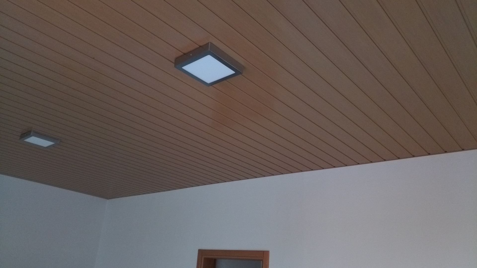 Teto de madeira com duas luminárias quadradas brancas acima de paredes brancas.
