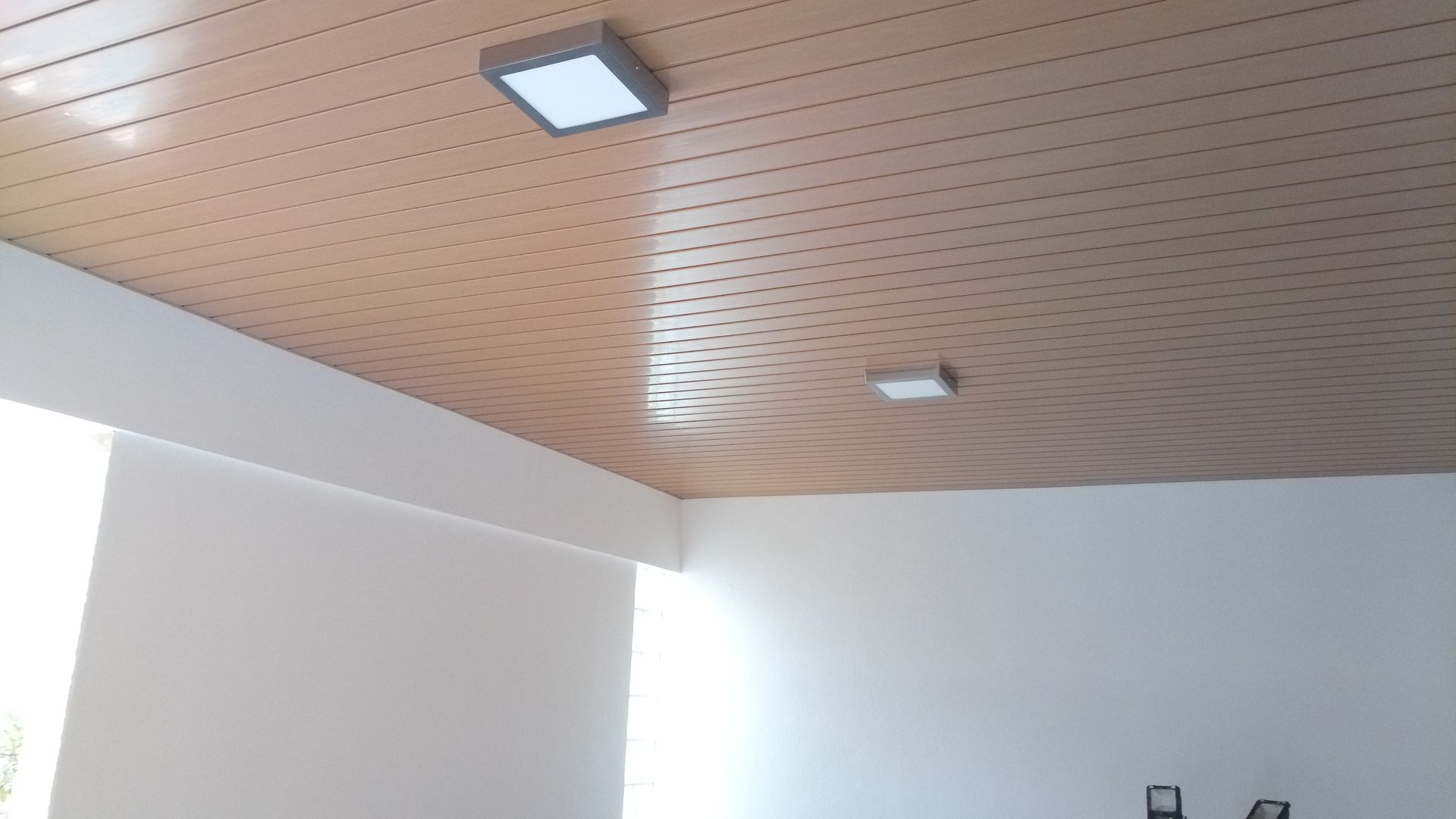 Teto perfurado em tom castanho claro com duas luminárias quadradas brancas em uma sala com paredes brancas.