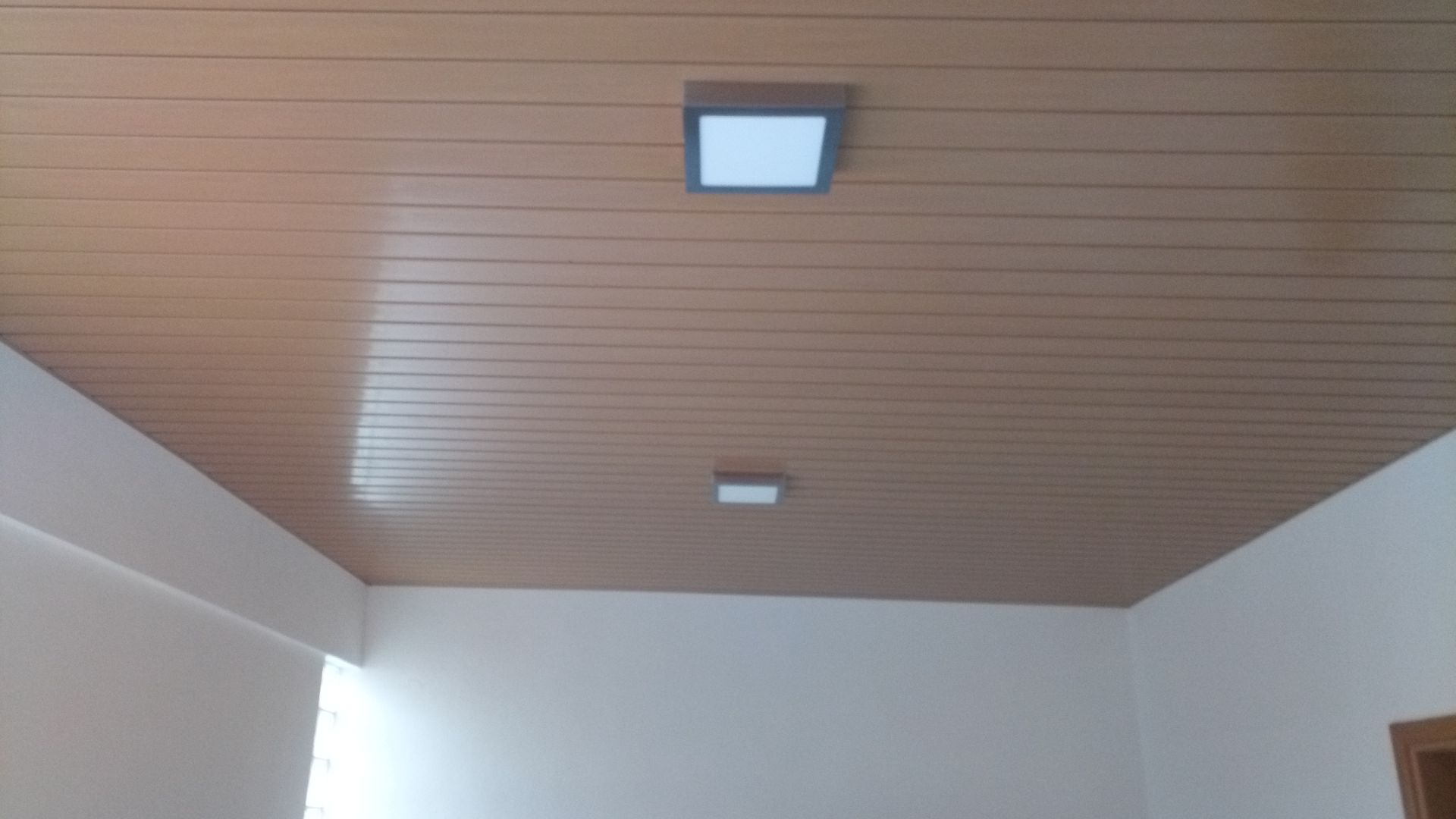 Teto com painéis marrons e duas luminárias quadradas com iluminação branca. Abaixo, paredes brancas.
