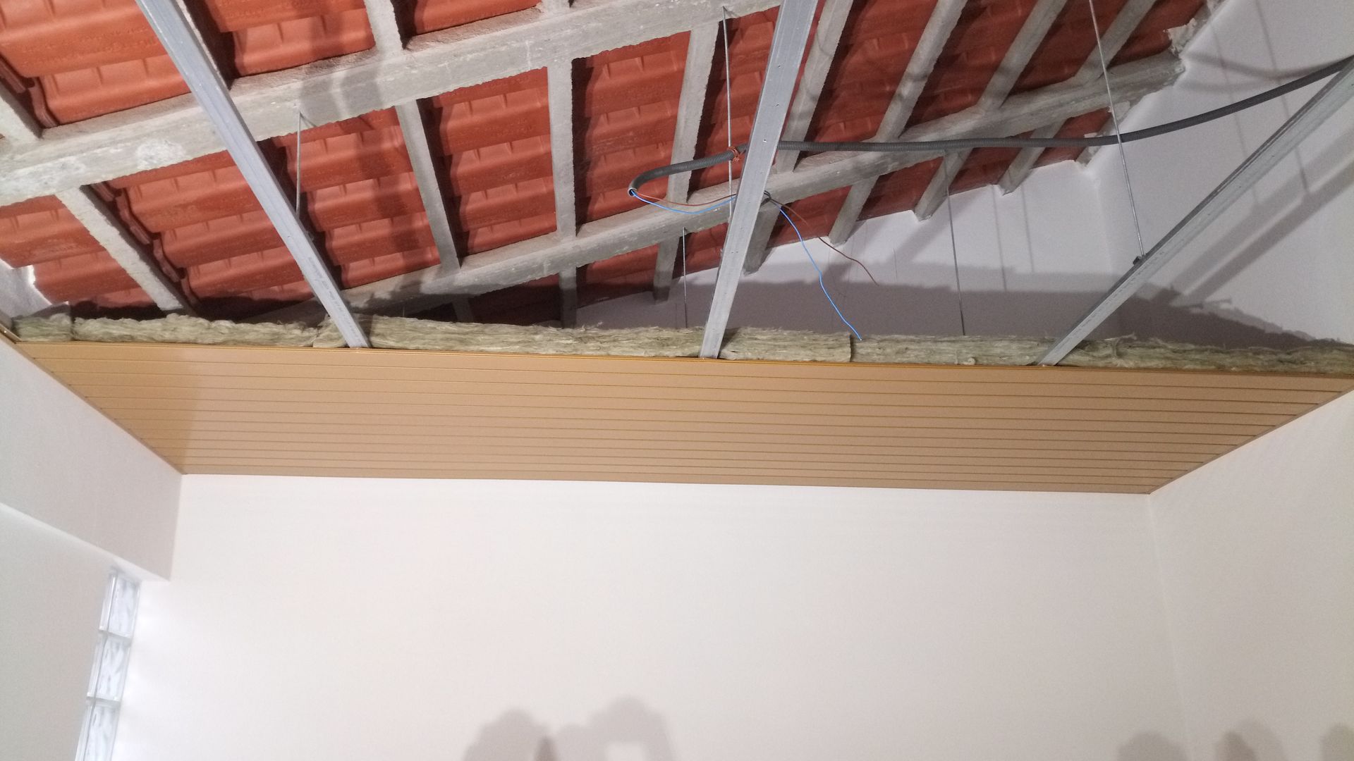 Teto em construção: painéis de madeira marrom, telhado de telha vermelha, paredes brancas.