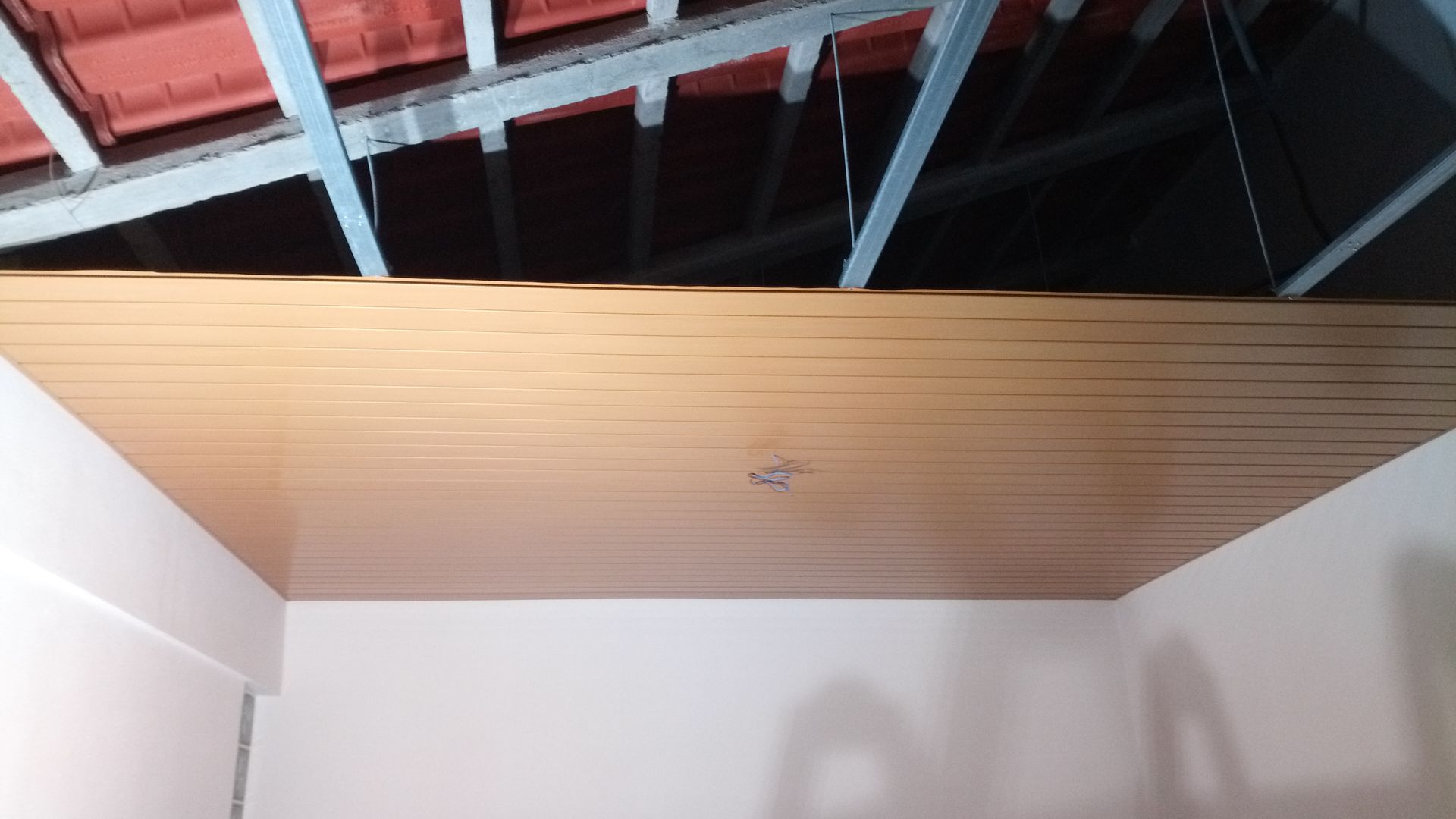 Painéis de madeira no teto, com vislumbre do telhado e suportes metálicos, fixados em paredes brancas.