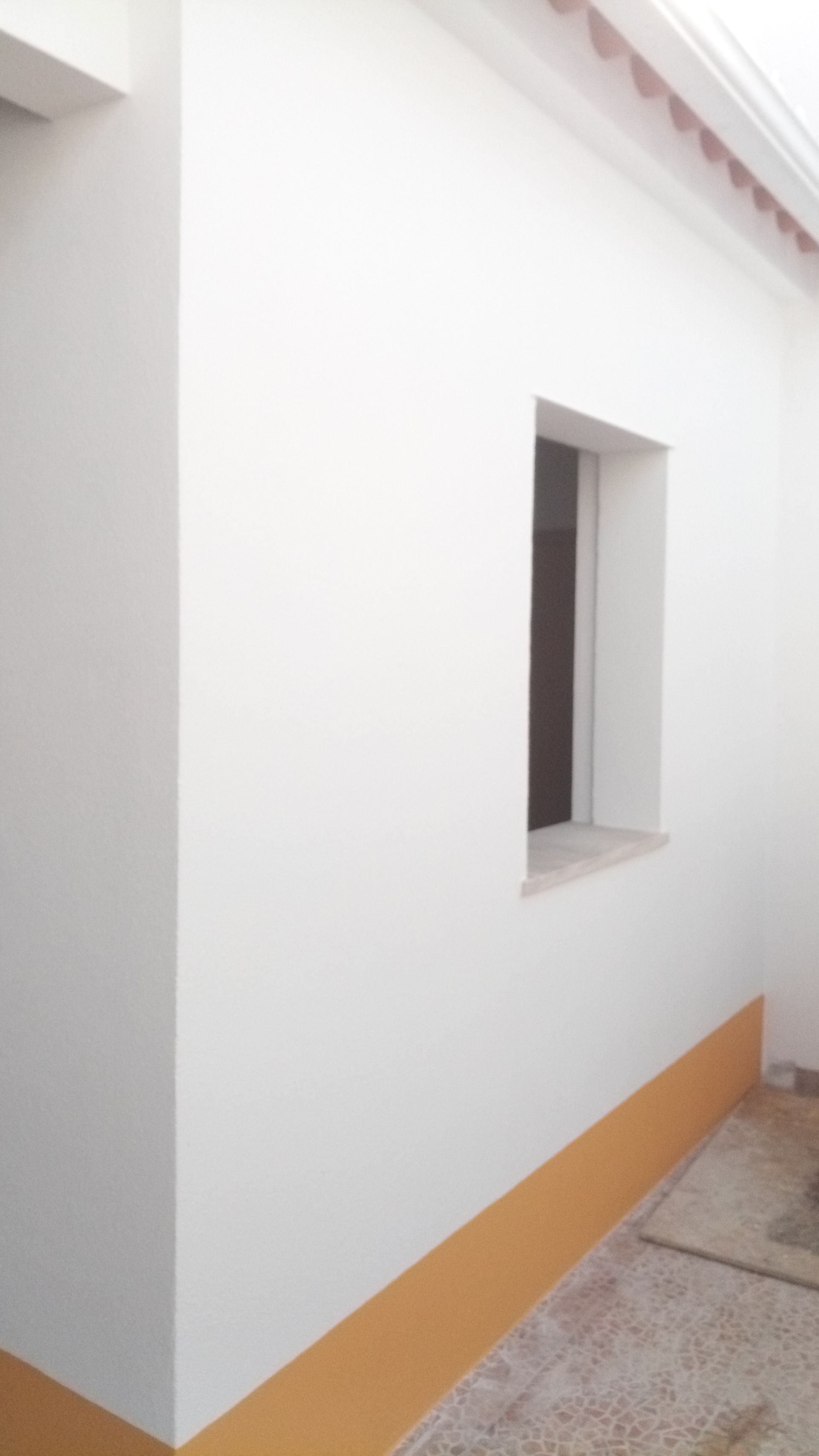 Exterior branco do edifício com uma pequena janela, detalhes em bege e beiral do telhado em terracota.