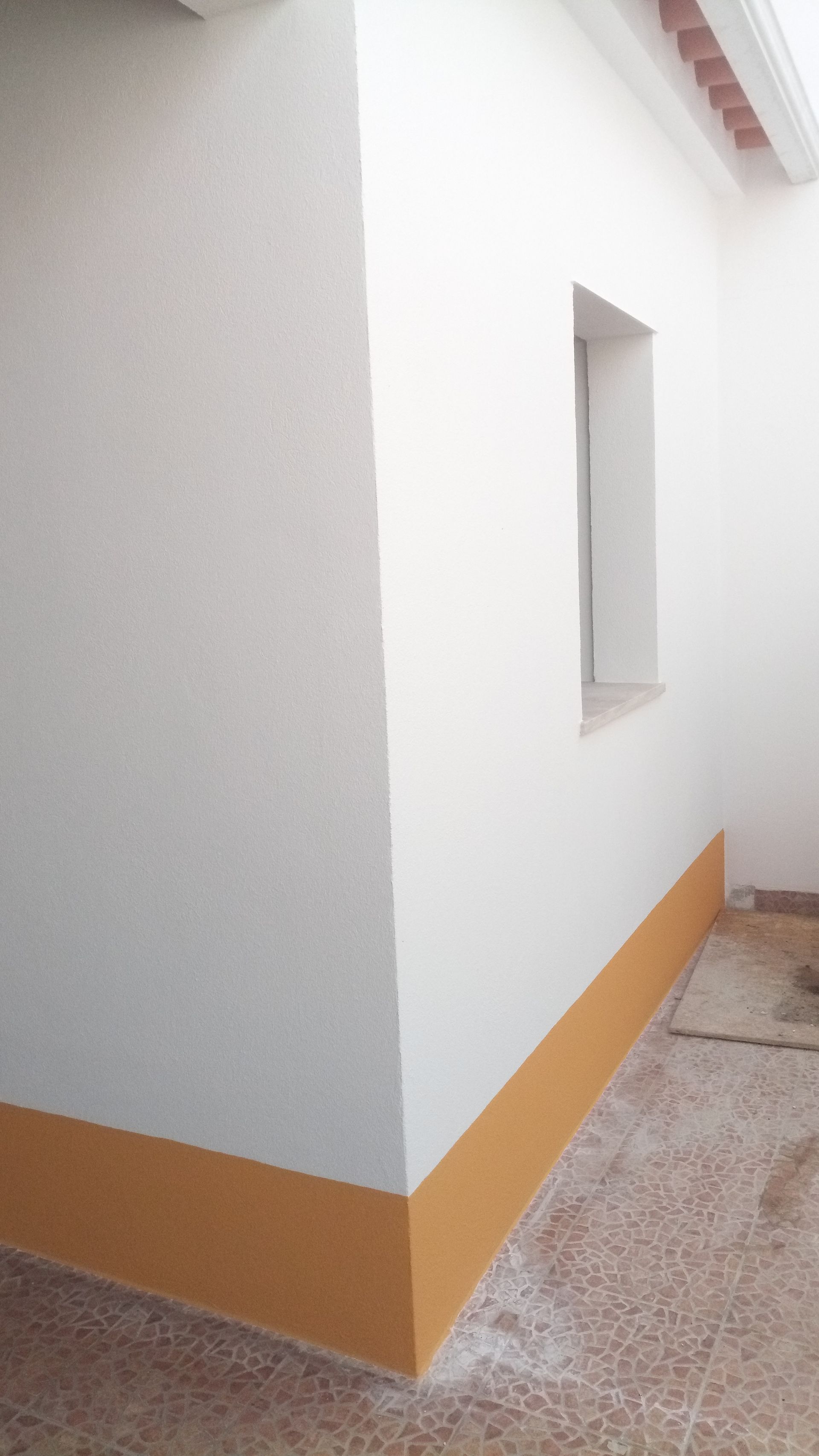 Canto de uma parede branca com rodapé laranja, uma pequena janela e piso salpicado.