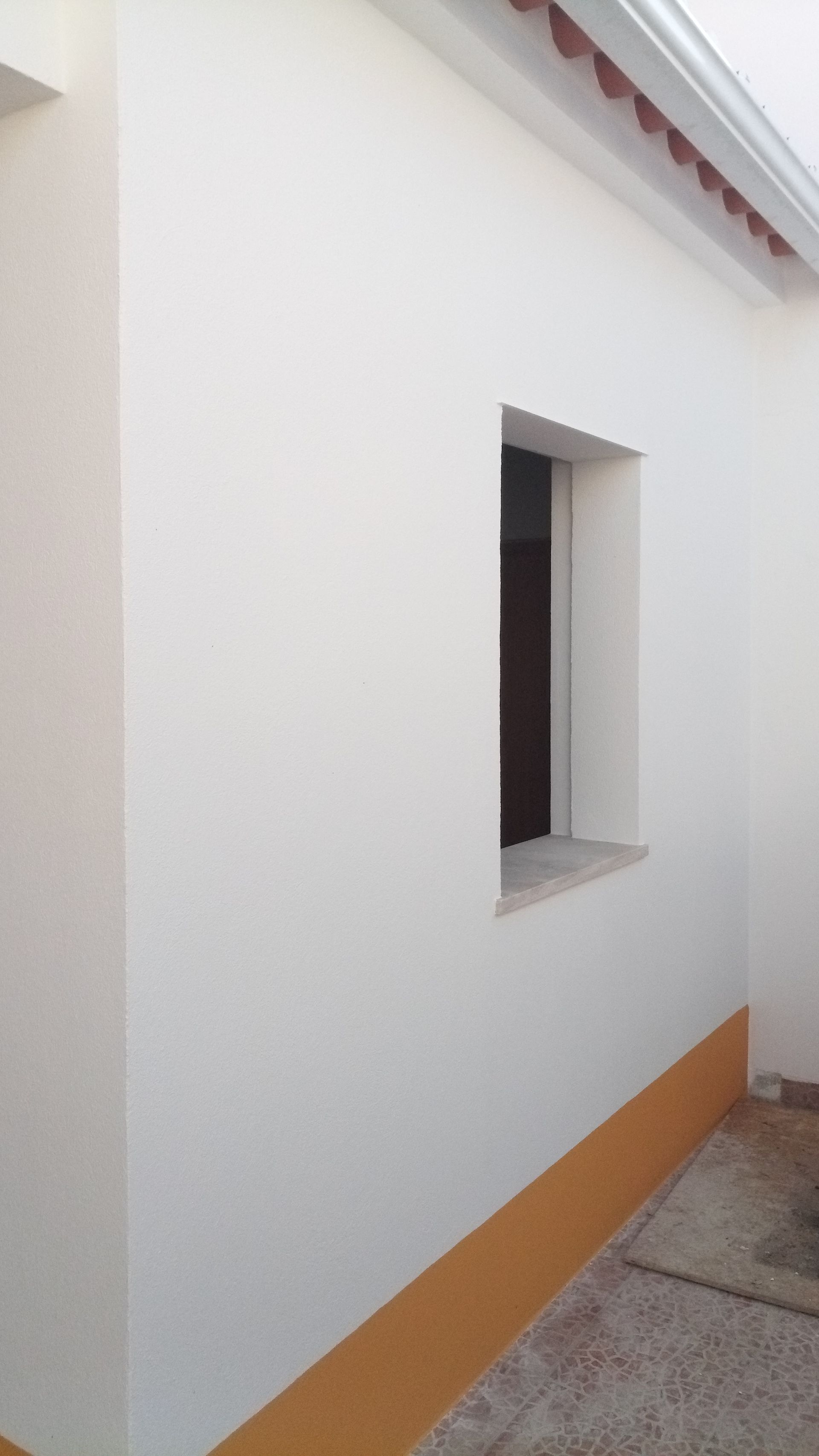 Exterior branco do edifício com uma pequena janela quadrada; faixa amarela na base.