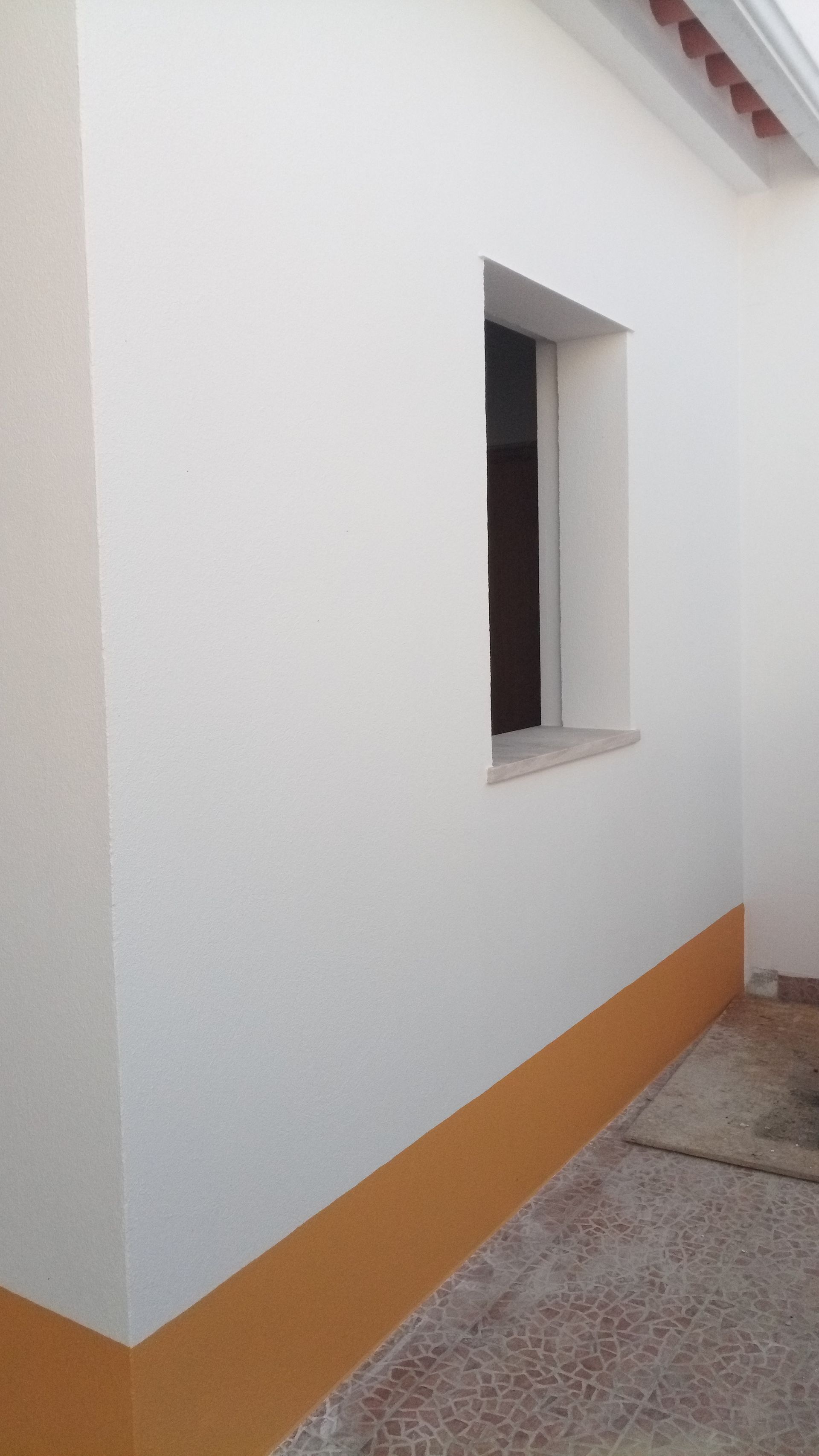 Exterior de prédio branco com uma pequena janela e uma faixa laranja próxima ao chão.