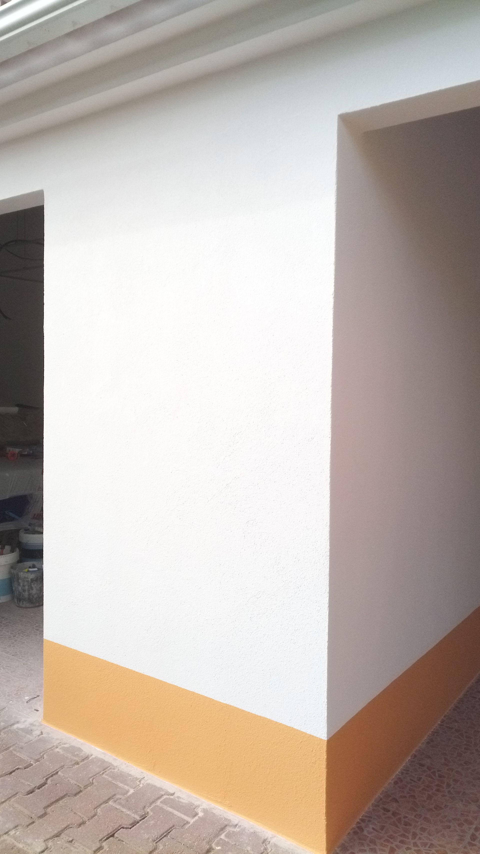 Canto de um edifício pintado de branco e amarelo, com porta de entrada, paredes texturizadas e piso de azulejo.