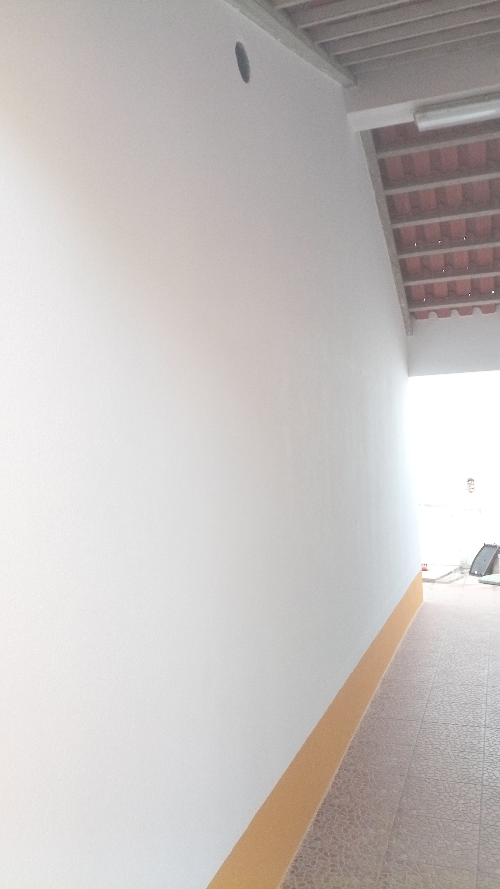 Parede branca com rodapé marrom, sob um telhado de telhas de terracota, com uma abertura circular preta no topo da parede.