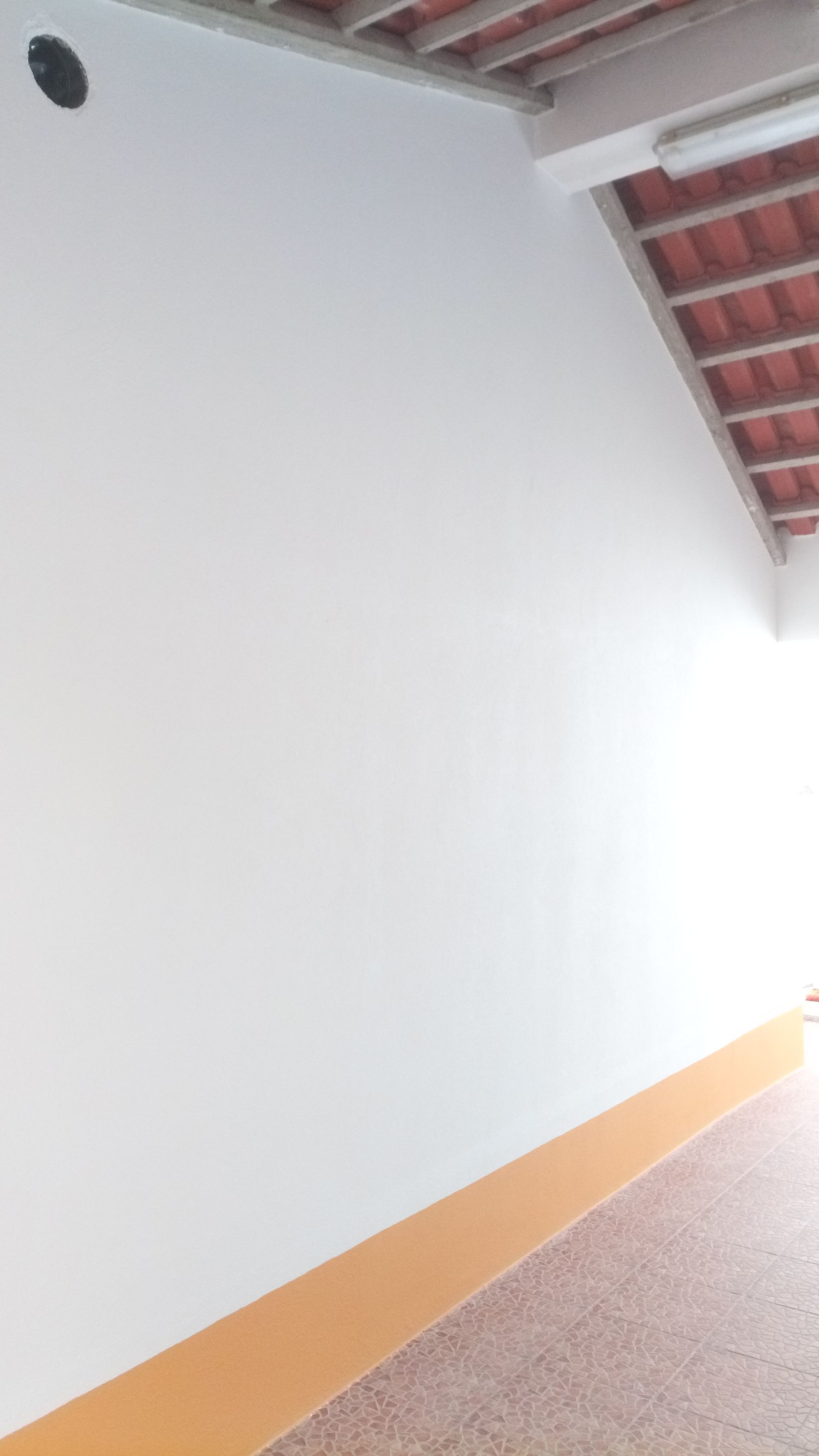 Parede branca com faixa laranja na parte inferior, corredor com teto de telha vermelha visível.