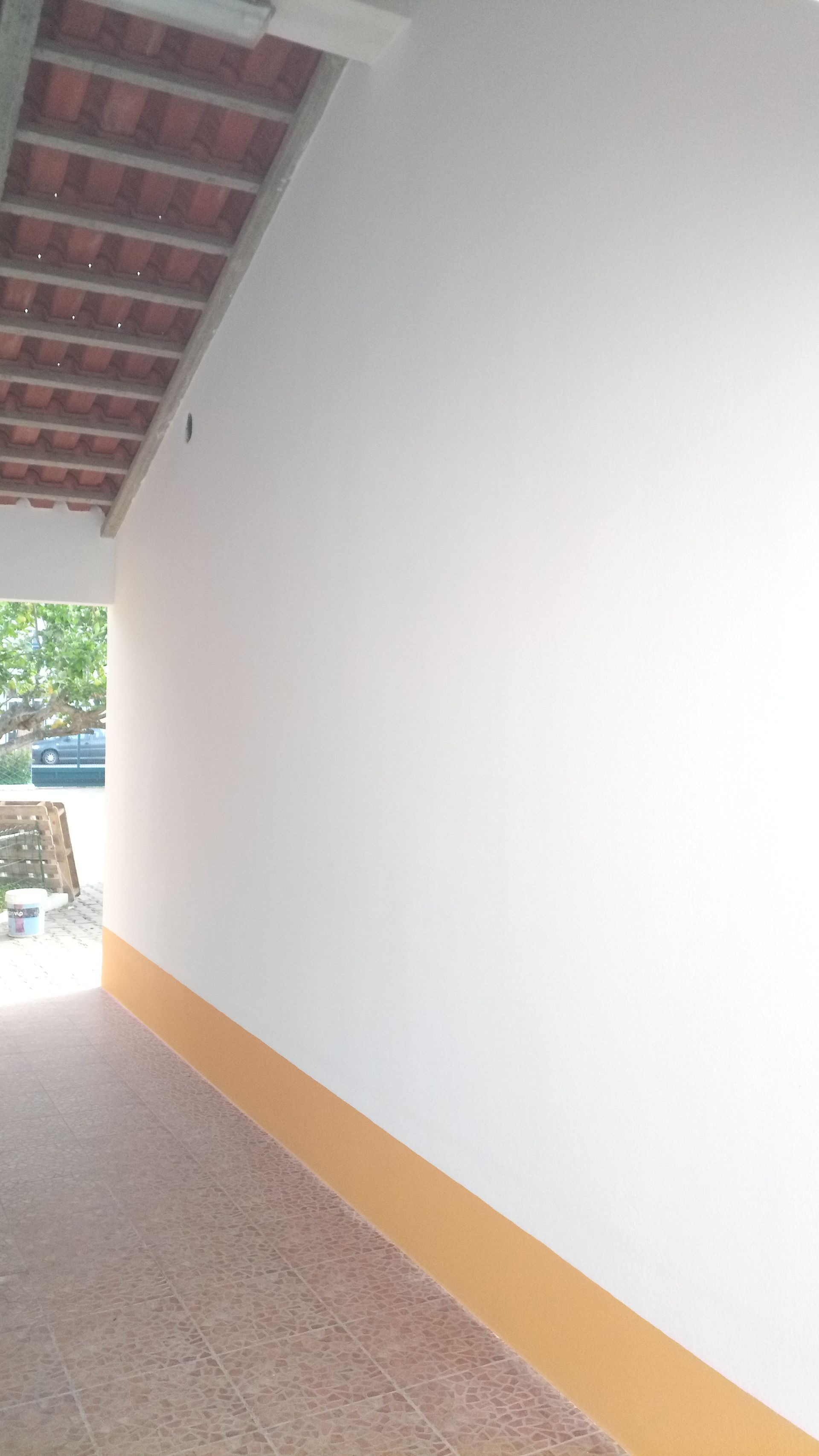 Parede branca com rodapé laranja, ao lado de uma passarela de azulejos sob um telhado de telhas vermelhas.