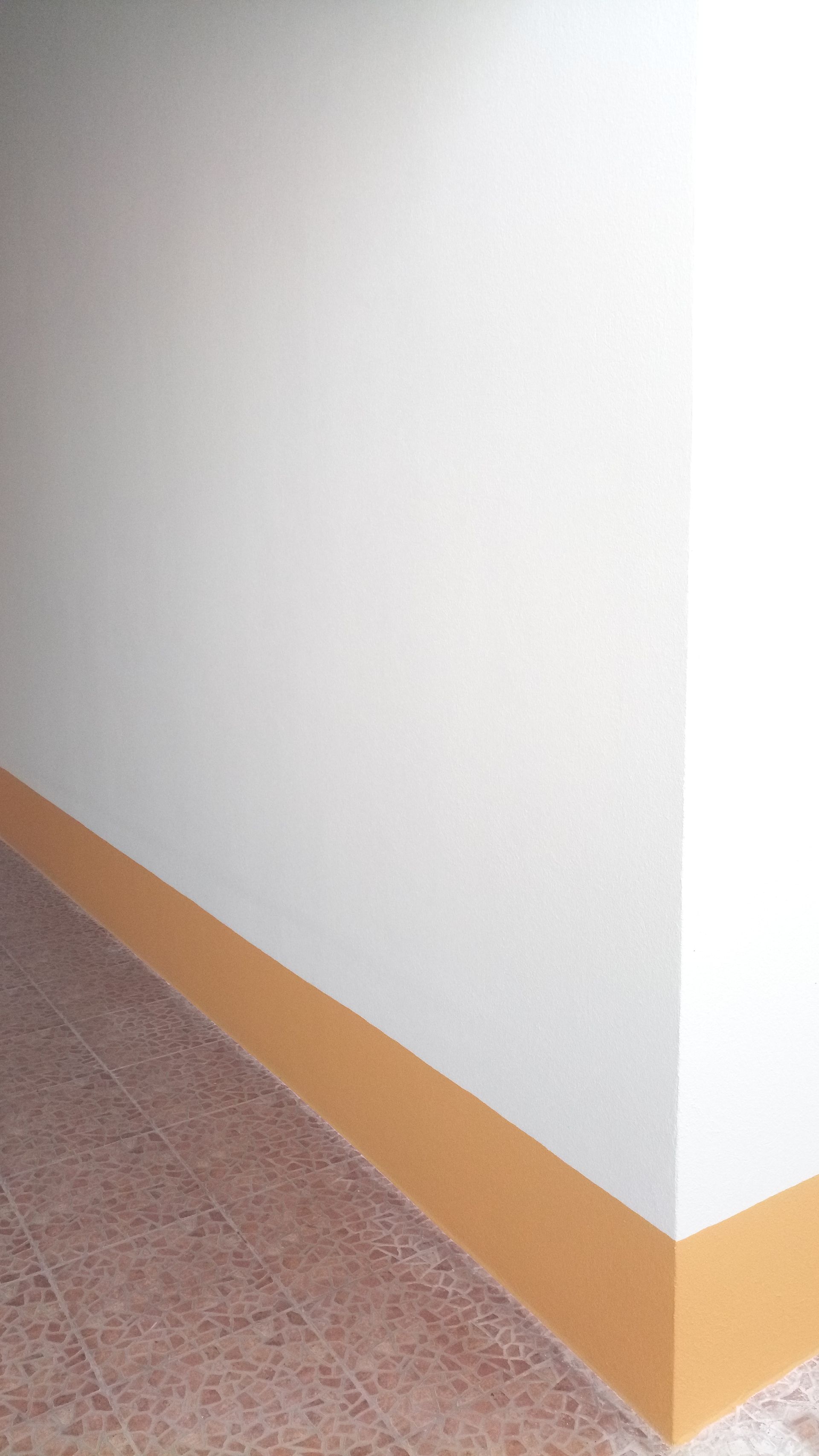 Canto de um quarto; paredes brancas, rodapé bege e ladrilhos marrons no chão.