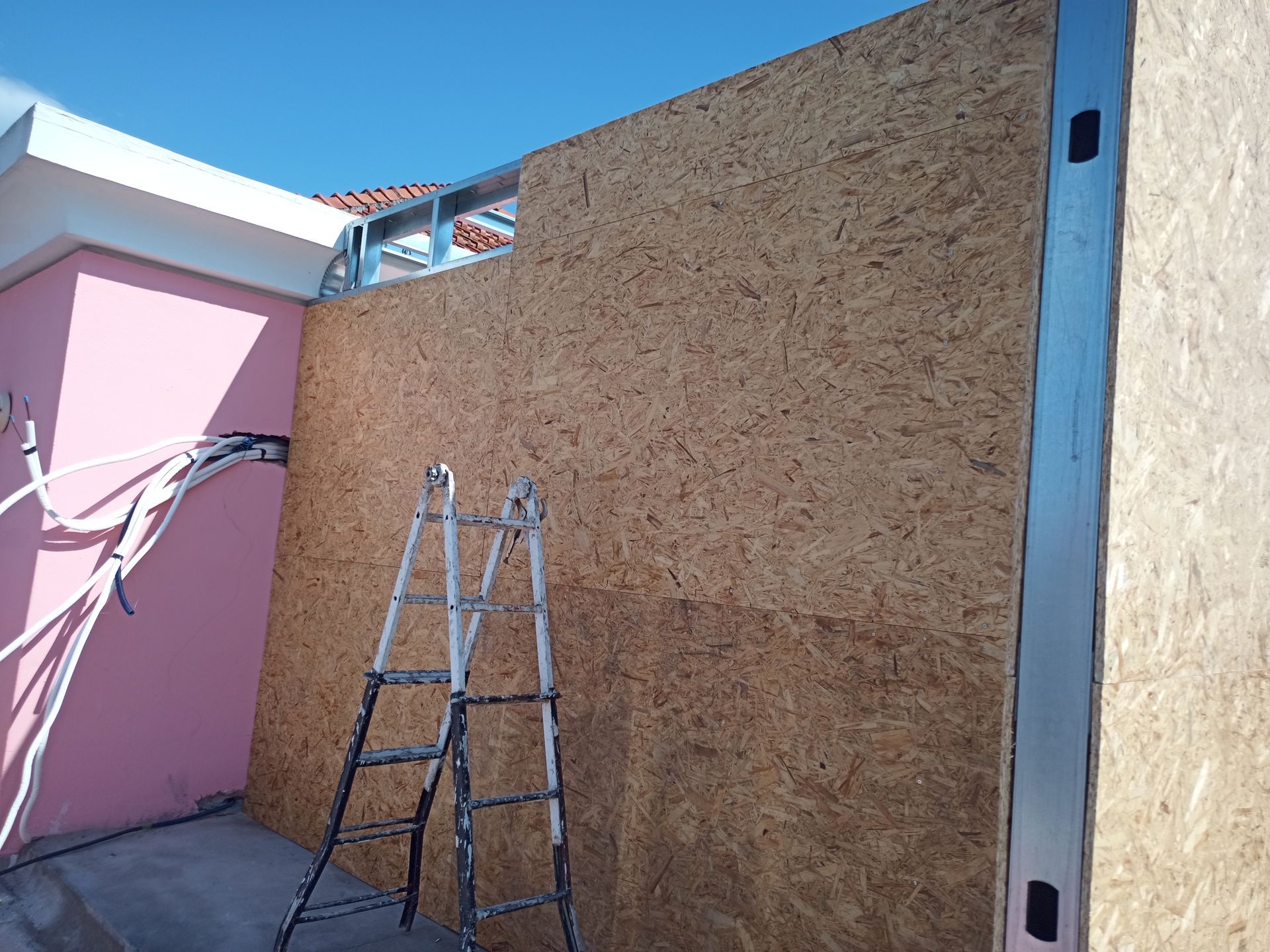 Construção de uma parede externa: painéis OSB sobre estrutura metálica, escada, prédio rosa, céu azul.