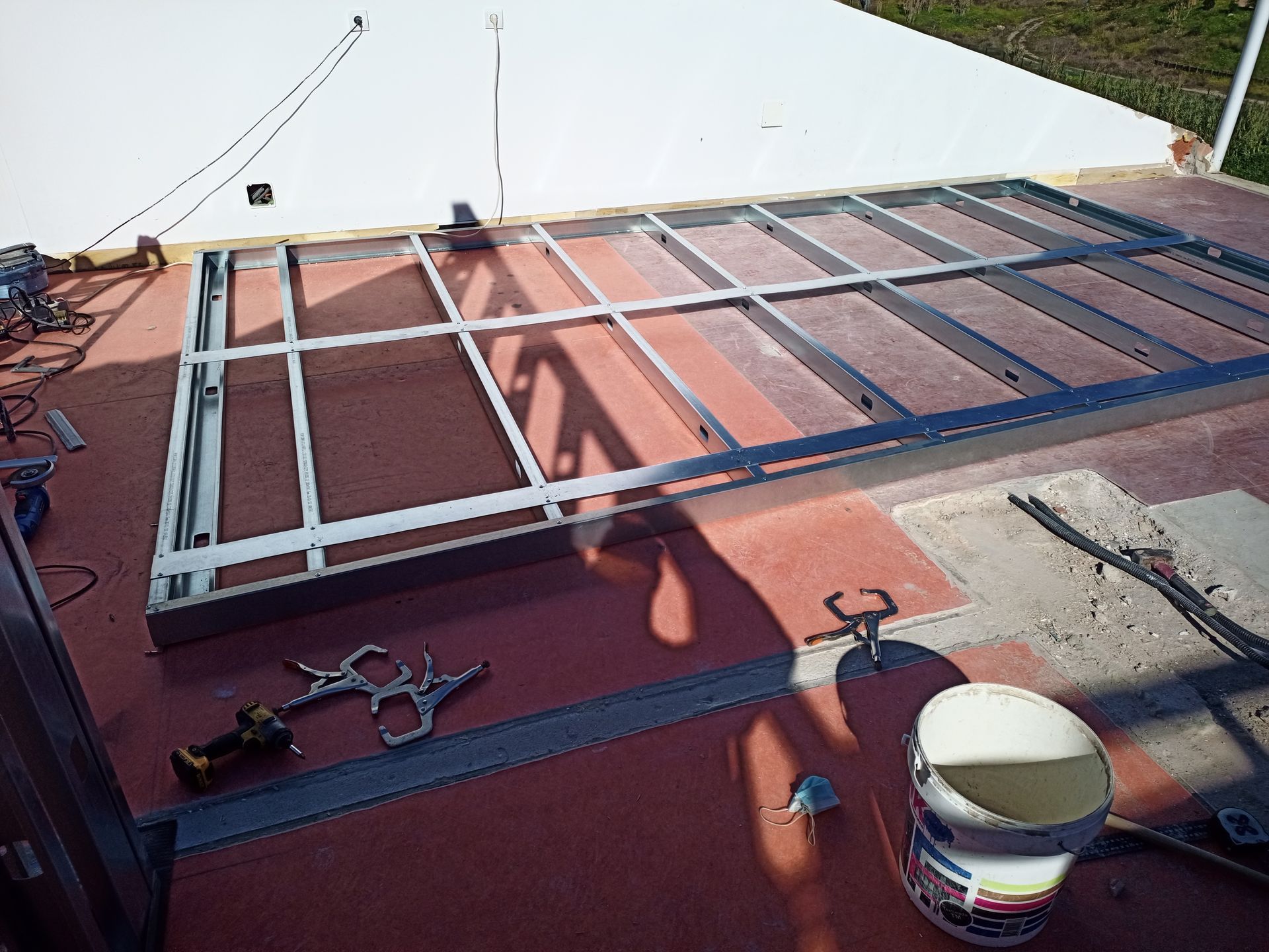 Estrutura metálica sobre uma superfície vermelha, com ferramentas e balde de tinta por perto.