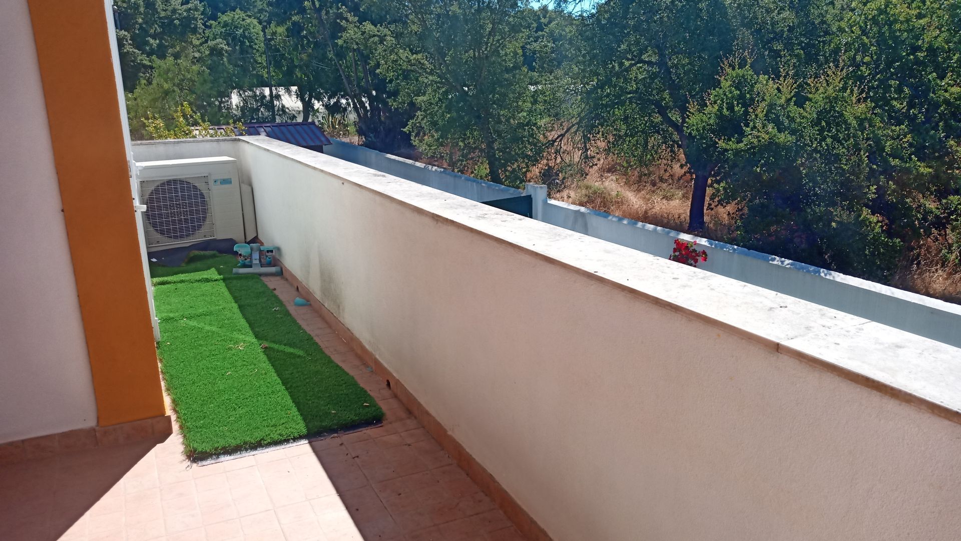 Varanda com grama artificial, ar condicionado e paredes brancas, com vista para as árvores.