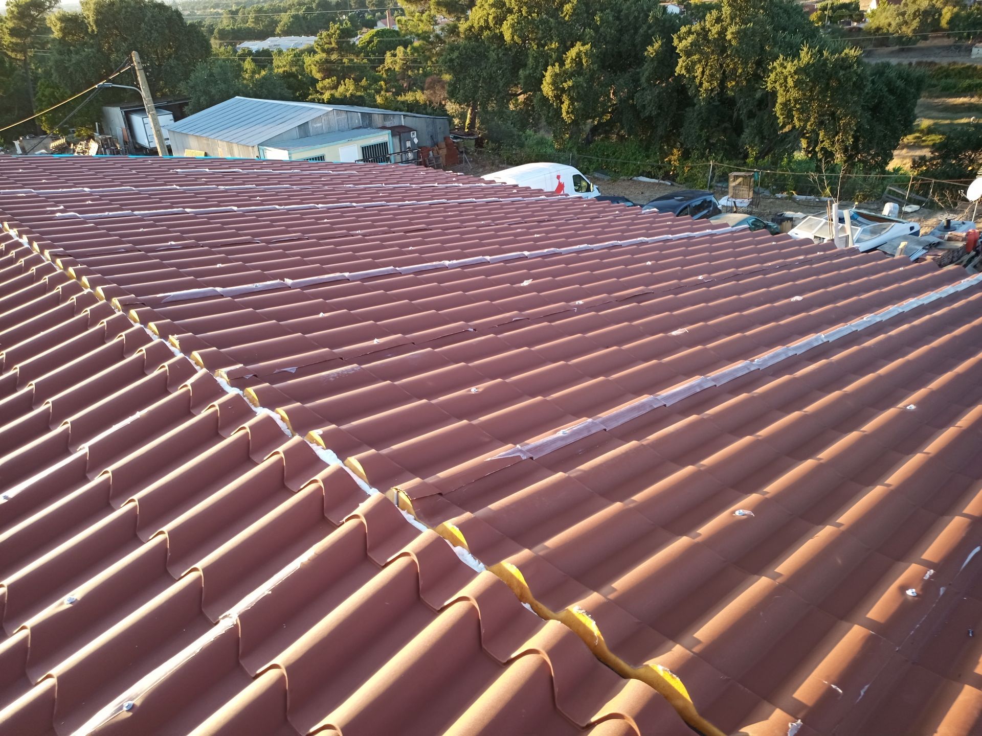 Telhado de zinco vermelho com luz solar, prédios e árvores ao fundo.
