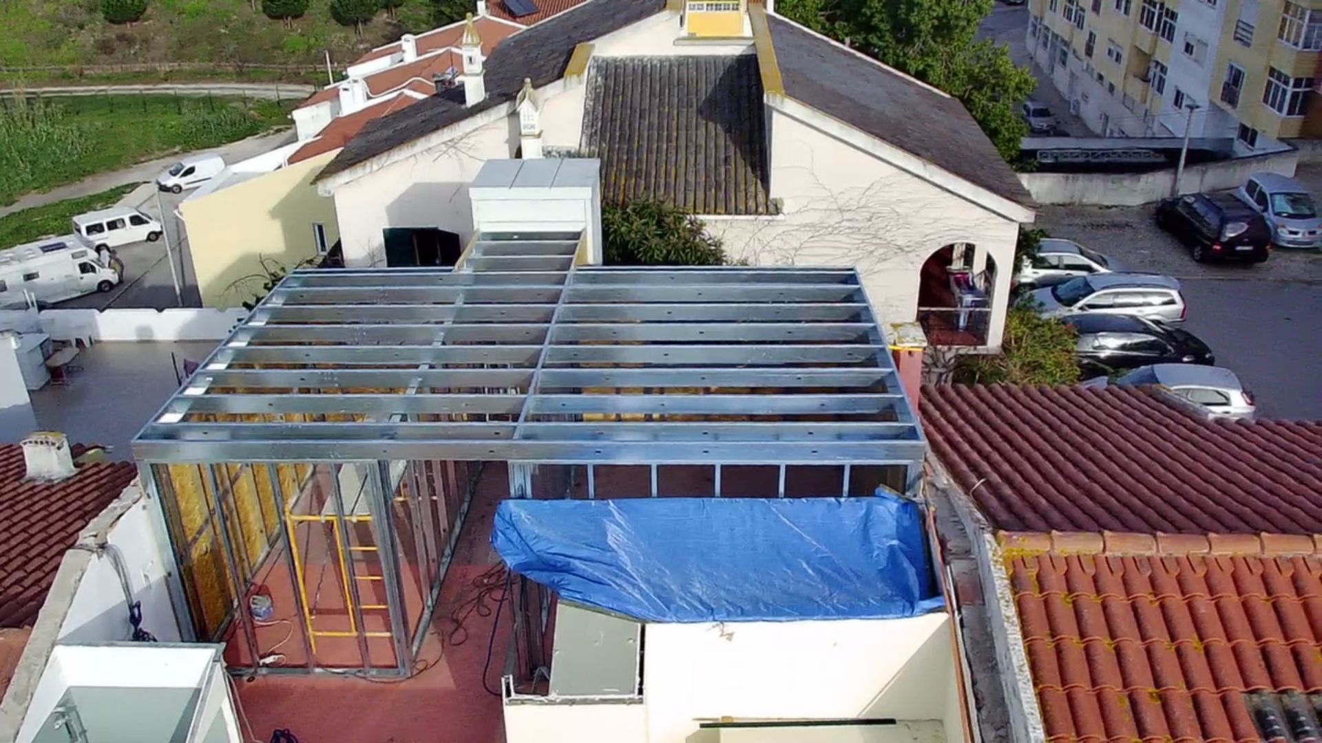 Canteiro de obras em um telhado, estrutura metálica, lona azul, prédios residenciais ao fundo.