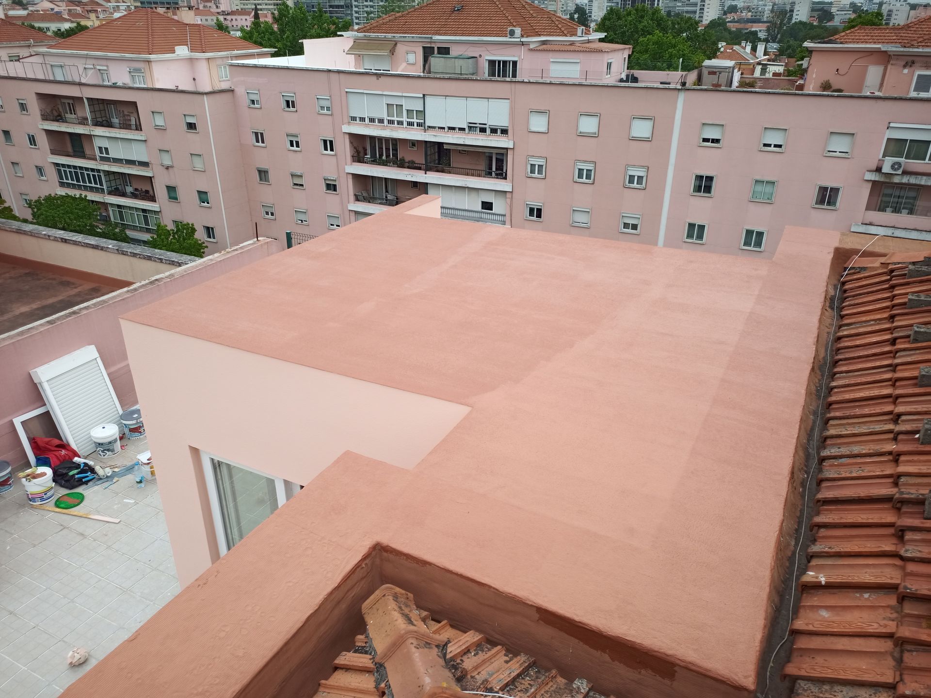 Vista do telhado de um prédio com telhas de terracota, ao lado de um telhado plano recém-pintado, com prédios cor-de-rosa ao fundo.